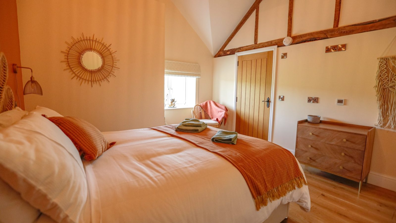 A cozy bedroom at Waen Farm House features a large bed adorned with orange and white linens and neatly folded towels. The room includes a wooden dresser, a sunburst mirror, exposed wooden beams, and a window with a woven blind. Soft, warm lighting enhances the inviting atmosphere.