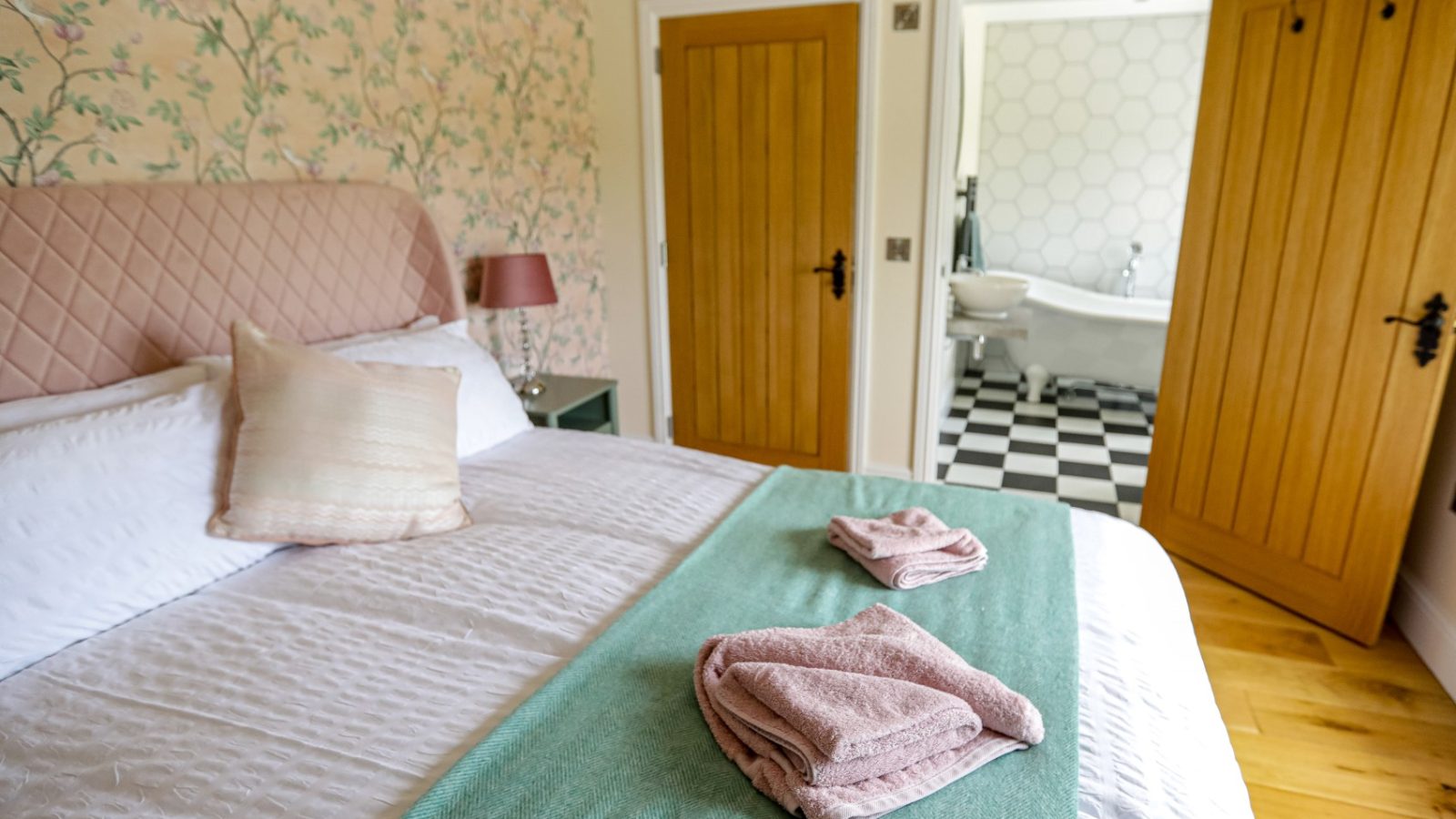 A cozy bedroom at Waen Farm House boasts a comfortable bed adorned with white and green bedding, complemented by two neatly folded pink towels. The room features floral wallpaper, a bedside table with a pink lamp, light wood doors, and an open door leading to a bathroom with a checkered floor and bathtub.