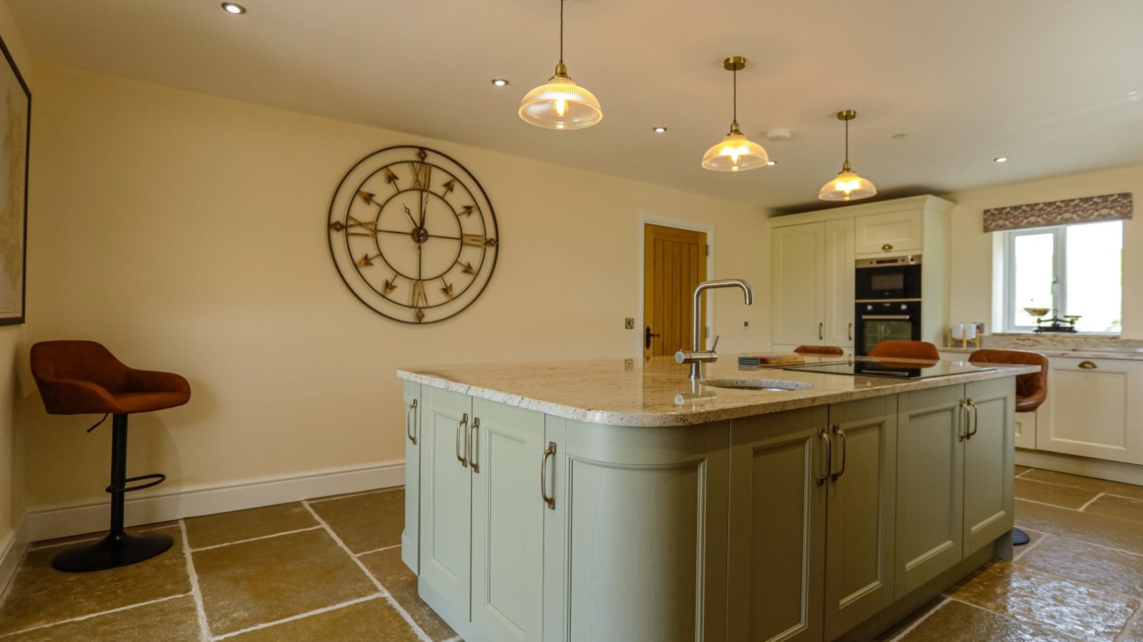 A spacious, modern kitchen at Waen Farm House features a large island with a sink, light-green cabinets, and a beige countertop. Three pendant lights hang above the island. The back wall has a circular wall clock, and there are bar stools next to the island. A large window lets in natural light.