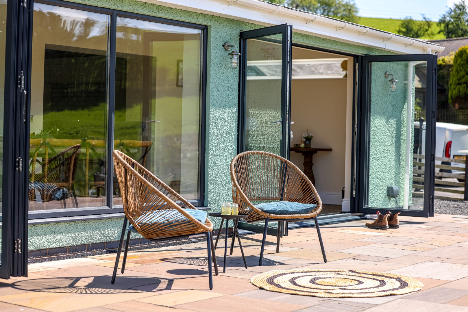 A patio featuring two wicker chairs with blue cushions, a small round table holding drinks, and an open door to the home beckons relaxation. The stone tiles host a round woven rug at the centre, while lush green scenery reflects in the large glass windows