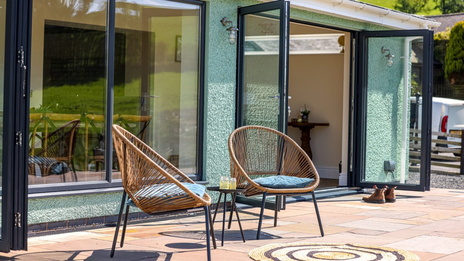 A cozy patio with two wicker chairs and cushions is positioned outside the Waen Farm House, featuring large glass doors. A round rug sits in front of the chairs, and a pair of shoes is nearby. The glass doors are open, revealing a wooden table inside.