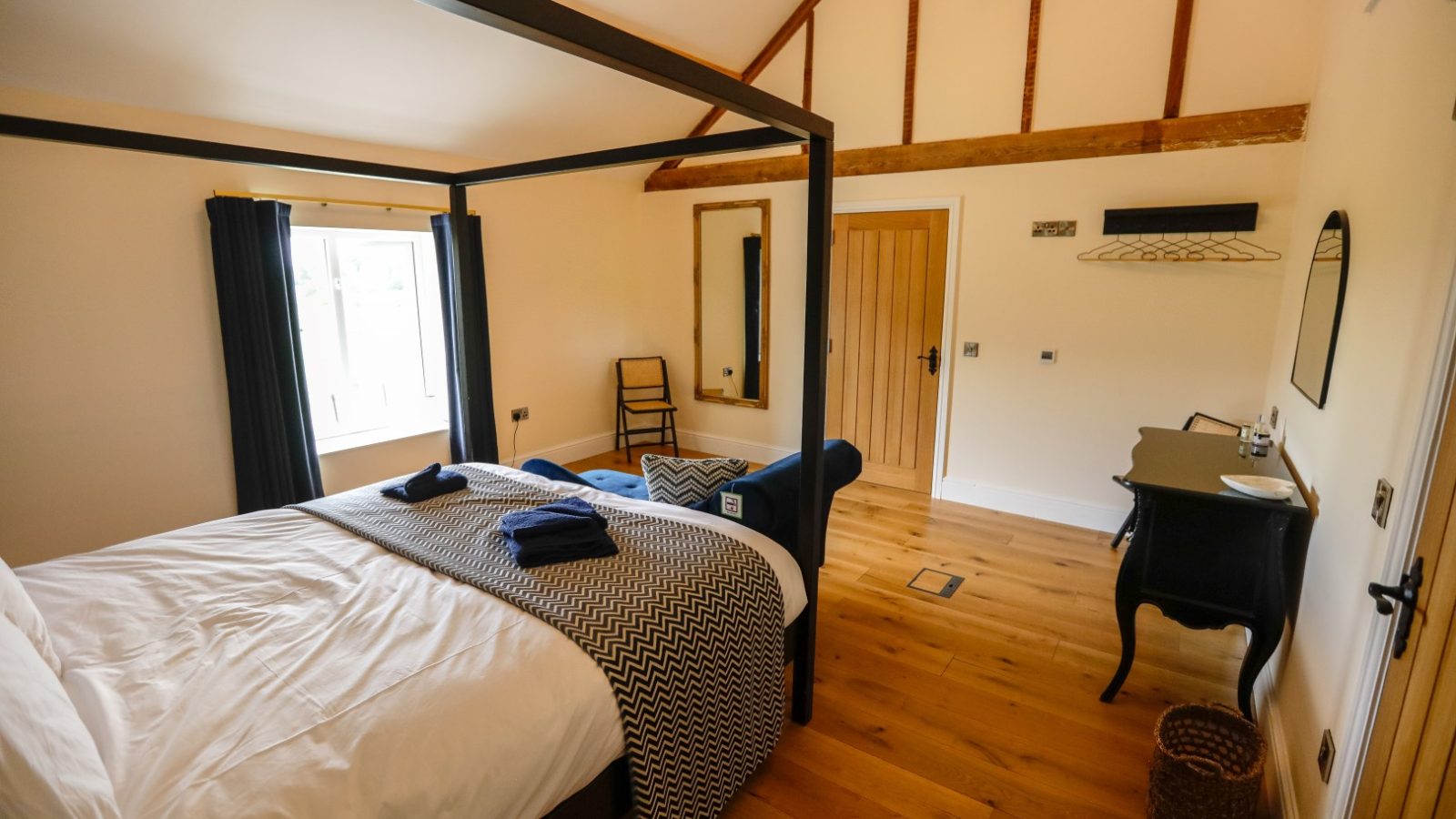 A cozy bedroom at Waen Farm House with wooden flooring features a double bed with a black frame and geometric-patterned bedding. The room includes a wooden chair, a small black desk with a round mirror, and a wicker basket. A window with dark curtains allows light into the space.