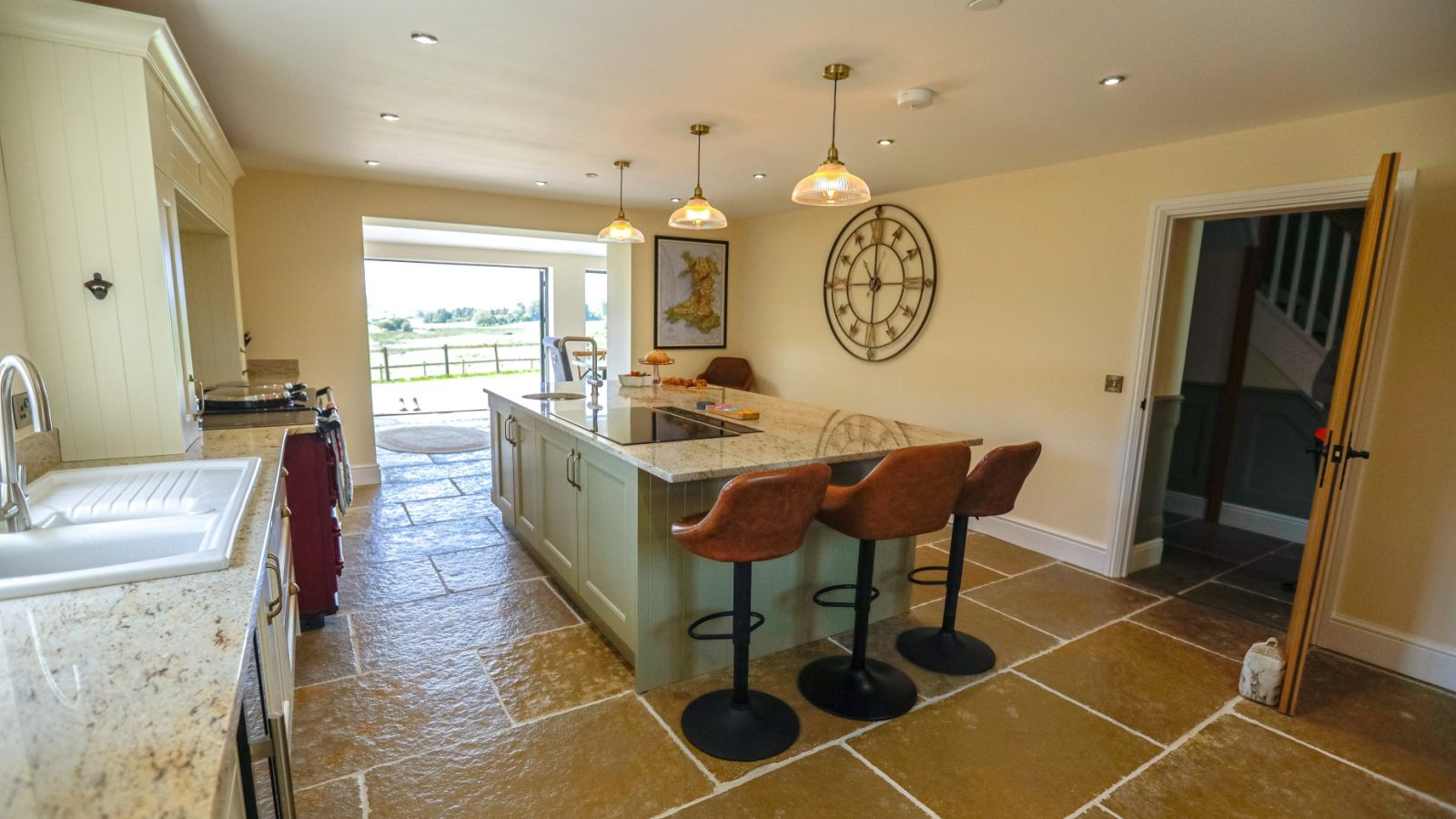 A spacious kitchen in the Waen Farm House with beige stone tile flooring features a long island with a marble countertop and three brown leather bar stools. Pendant lights hang above the island, there's a large clock on the wall, and an open door reveals a view of a green landscape.