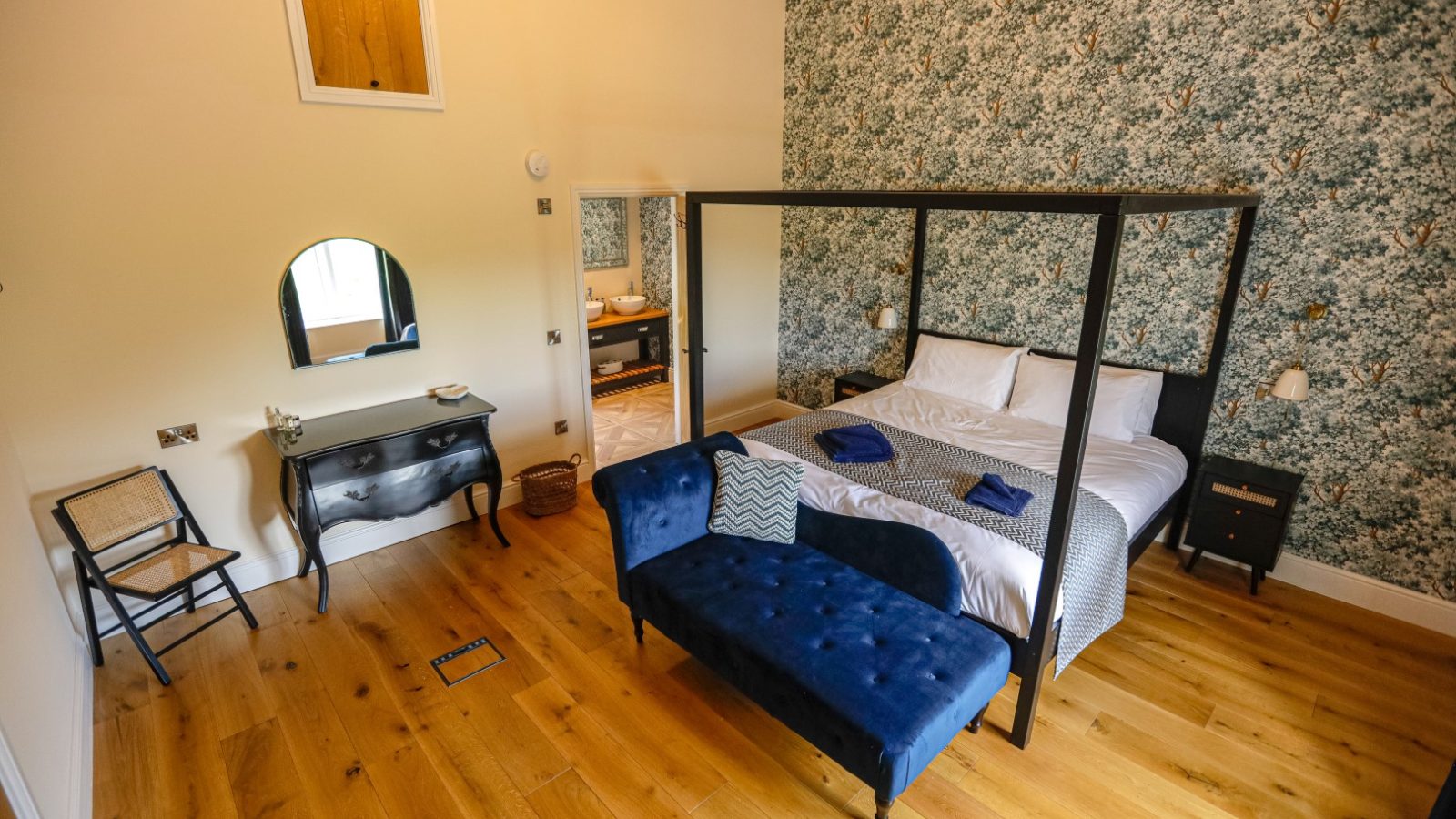 A cozy farmhouse bedroom with wooden flooring features a four-poster bed with white and blue linens. A blue tufted bench sits at the foot of the bed. A wooden dresser with a round mirror and a wicker chair are placed against the floral-patterned wallpapered walls, creating a charming rustic vibe.