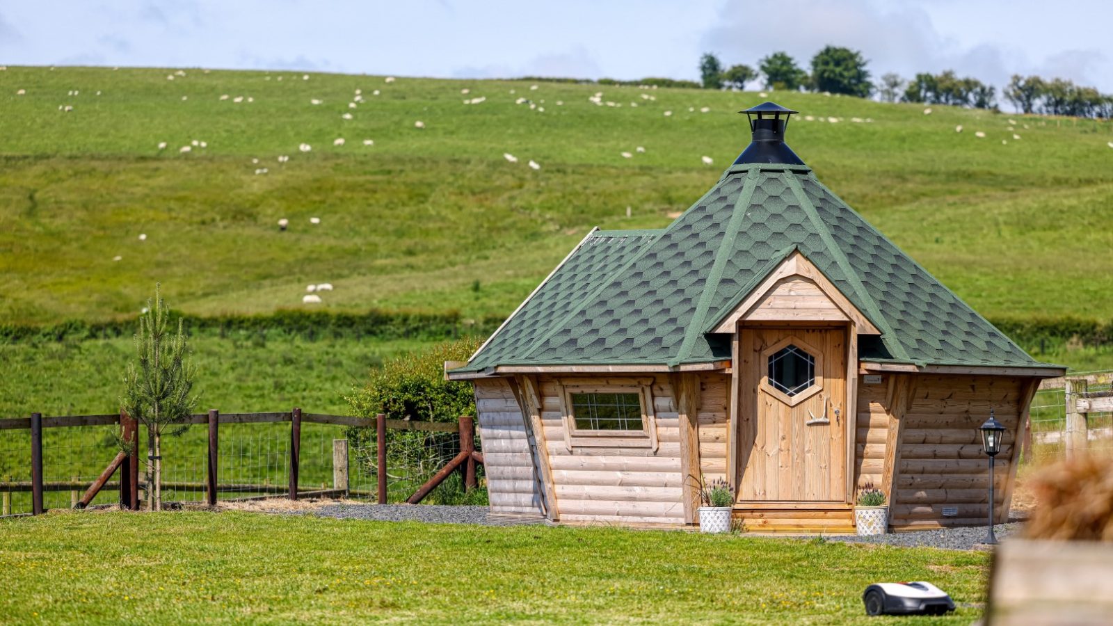 A quaint, small wooden cabin with a hexagonal shape and a green shingled roof sits in the lush landscape of Waen Farm House. The cabin has a distinctive chimney and a hexagonal window on the wooden door. A fenced area and gentle, rolling hills with grazing sheep are in the background.