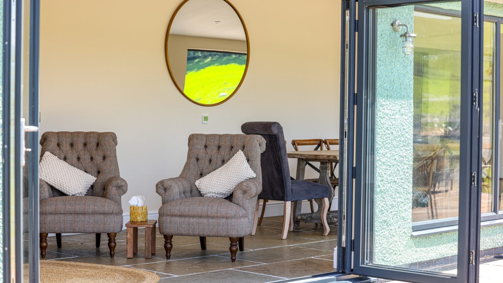 A cozy living area with two tufted armchairs and a round mirror on the wall. The room, evoking a charming farmhouse feel, features large glass doors that open to an outdoor space, allowing natural light to flood in. A small table with a box of tissues sits between the armchairs.