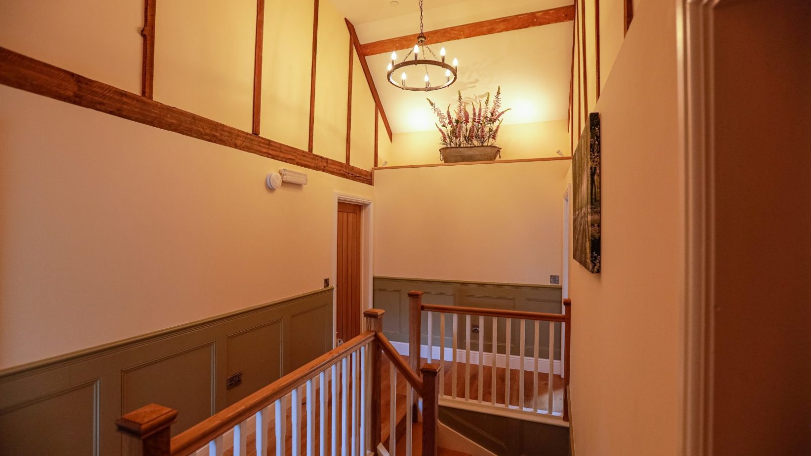 A warm, well-lit hallway with wooden beams and wainscoting embodies a charming farmhouse feel. Decor includes a circular chandelier and a floral arrangement atop a shelf. Wooden stairs with white railings lead to a landing, with doors visible in the background. Walls are painted in neutral tones.