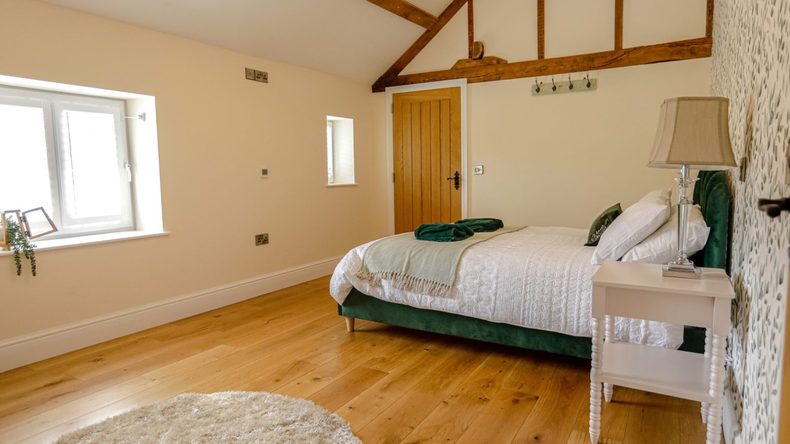 A cozy bedroom at Waen Farm House with a wooden floor, featuring a bed with white bedding and green accents. The room has a slanted wooden beam ceiling, a small nightstand with a lamp, a wooden door, and two windows allowing natural light to fill the space. A round rug is on the floor.