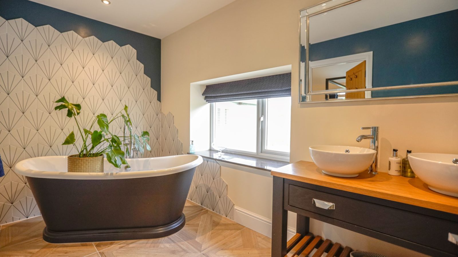 A modern bathroom in the Waen Farm House features a freestanding black bathtub with a potted plant on the edge. The wall behind the tub boasts an elegant geometric accent. A wooden vanity with double sinks and a large mirror is positioned beside a window with a dark shade.