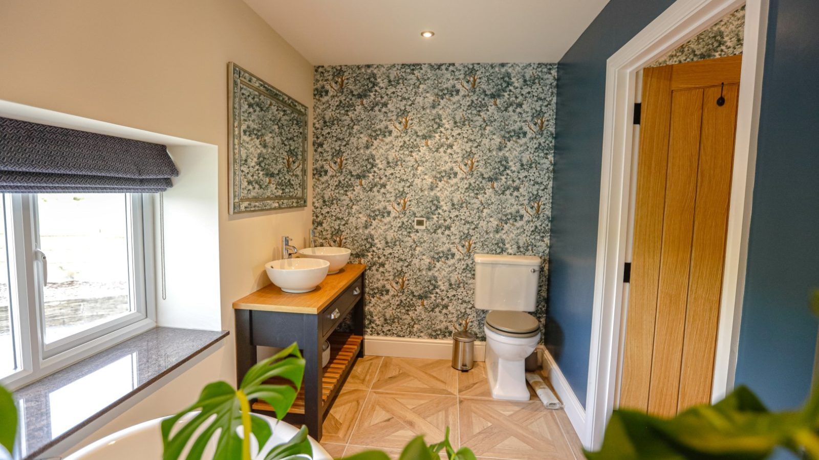 A modern bathroom featuring a toilet, a vanity with two vessel sinks, a large mirror, and a floral-patterned accent wall. The room also has a window with a dark roller shade, potted plant, and wooden flooring with a geometric design that evokes the charm of Waen Farm.