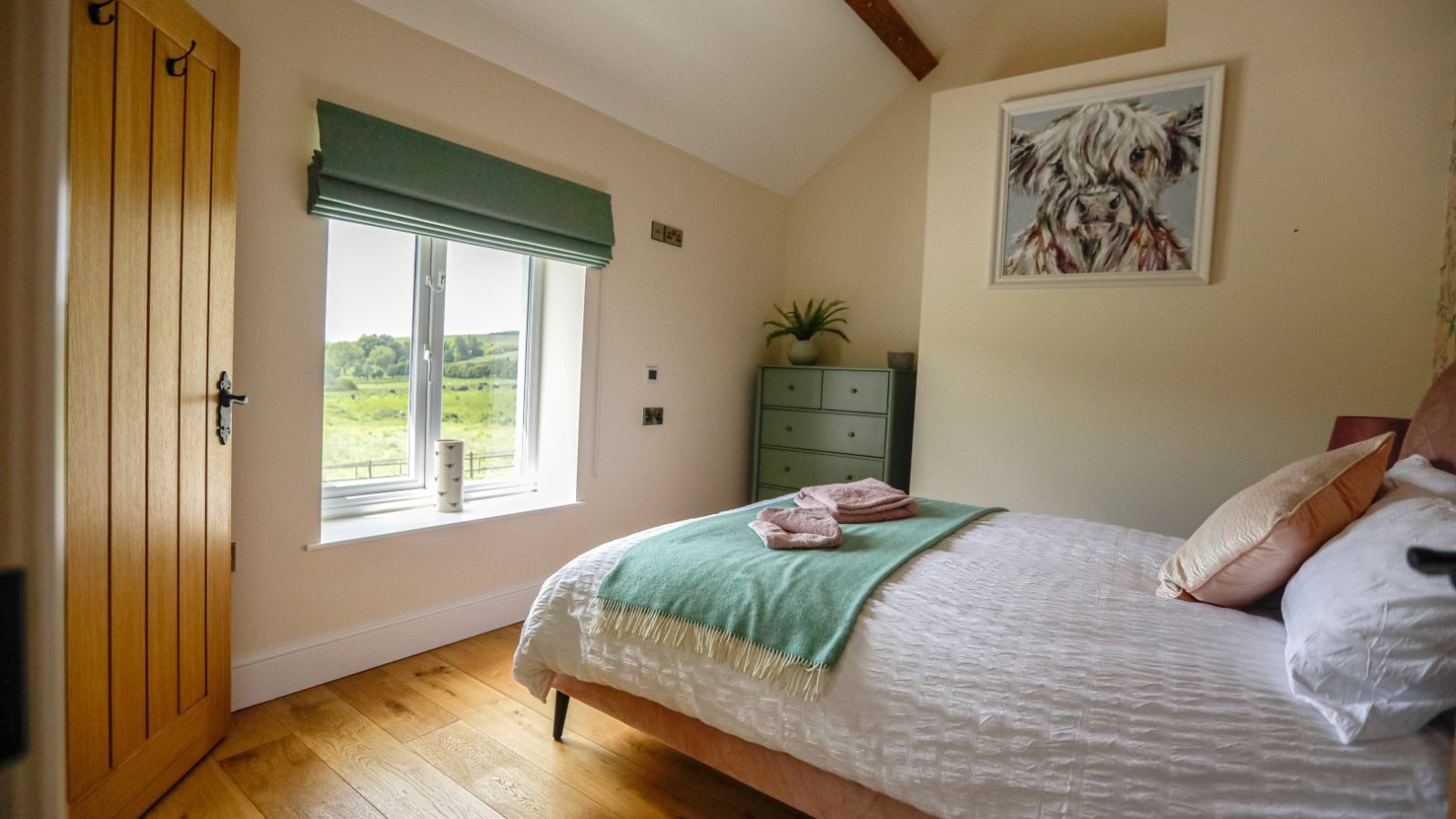 A cozy bedroom at Waen Farm House features wooden floors and a vaulted ceiling. A double bed with white linens, green and pink towels, and decorative pillows sits beside a window with a green Roman shade. A dresser and a framed picture of a highland cow adorn the wall. A lush landscape is visible outside.