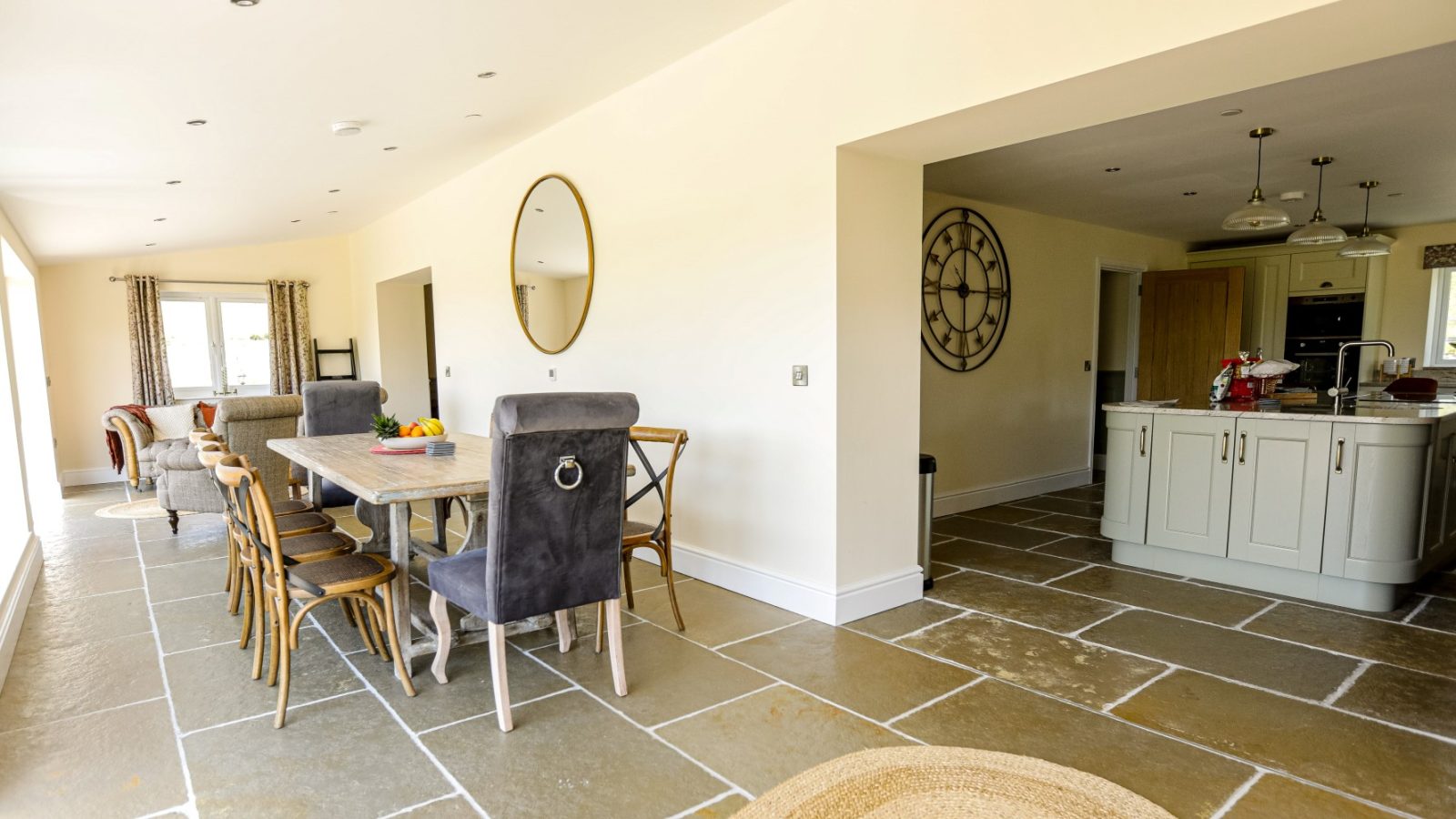 A modern kitchen and dining area reminiscent of the Waen Farm House, featuring beige walls and stone tile floors. The dining table is surrounded by a mix of wooden and upholstered chairs. Light cabinetry and stainless steel appliances adorn the kitchen, while a circular mirror and large wall clock decorate the walls.