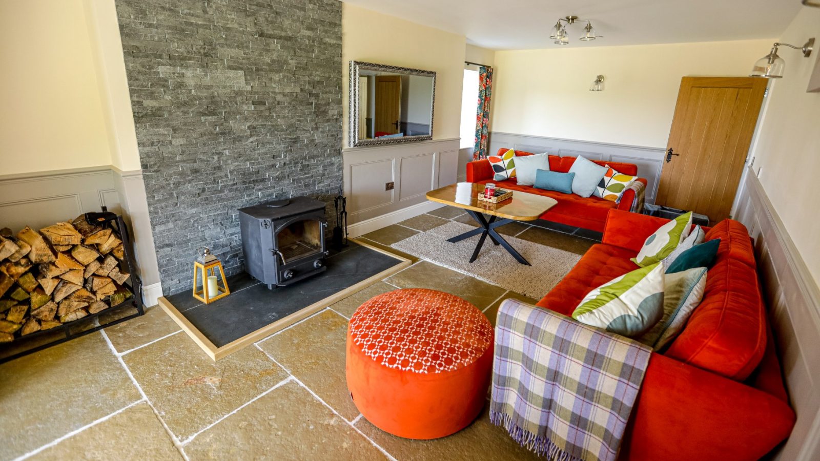 A cozy living room at Waen Farm House features a bright orange sectional sofa with colorful cushions, a round orange ottoman, a checkered throw, and a wooden coffee table. A wood-burning stove sits next to a stack of firewood on a stone hearth against a grey accent wall.