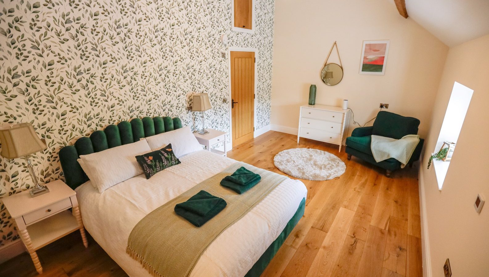 A bedroom at Waen Farm House boasts a green and white leaf-patterned accent wall, featuring a bed with a green headboard and neatly folded green towels. The room has a wooden floor, white furniture, a round rug, and a green armchair by the window. A mirror and painting adorn the walls.