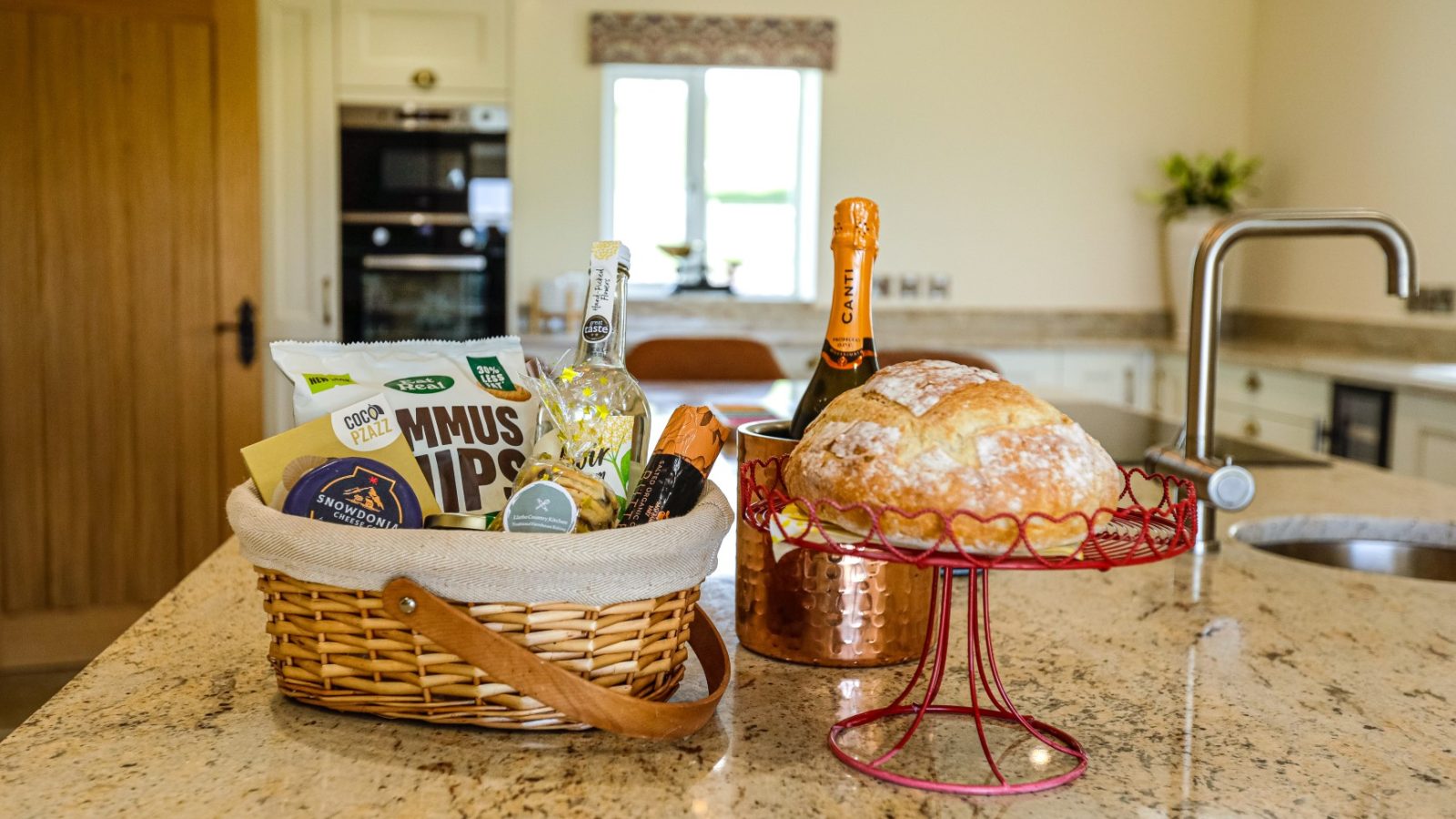 A kitchen counter at the Waen Farm House displays a wicker basket filled with assorted snacks and drinks, including a bag of chips and a bottle of soda. Next to the basket, a loaf of bread on a decorative red stand and a bottle of champagne in an ice bucket are also visible.