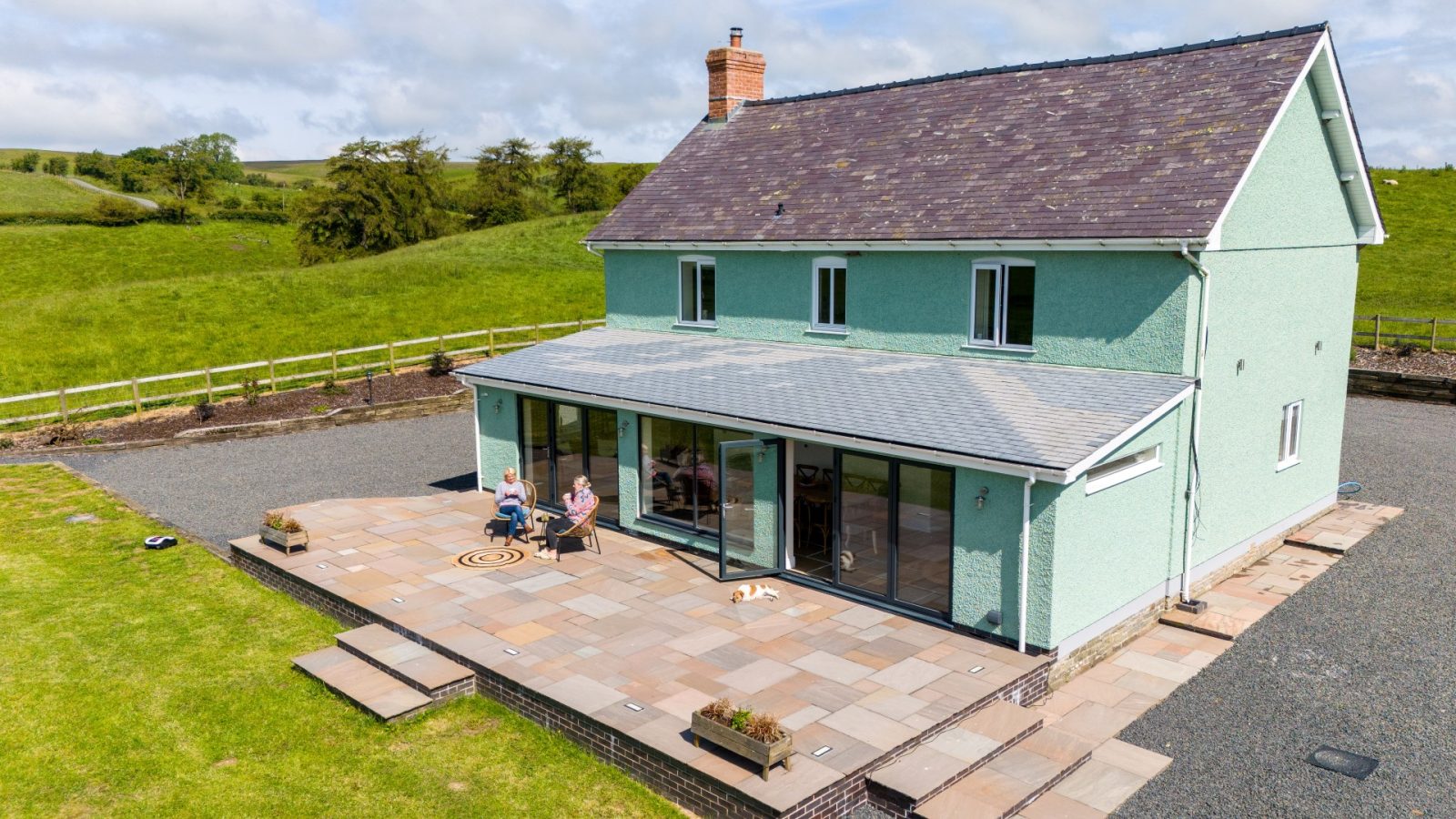 Aerial view of a light green two-story house with a sloped roof and large patio at Waen Farm. The patio has several potted plants, a table, and chairs. Two people are seated at the table, while a dog lies on the patio. The farm house is surrounded by a large grassy yard and countryside landscape.