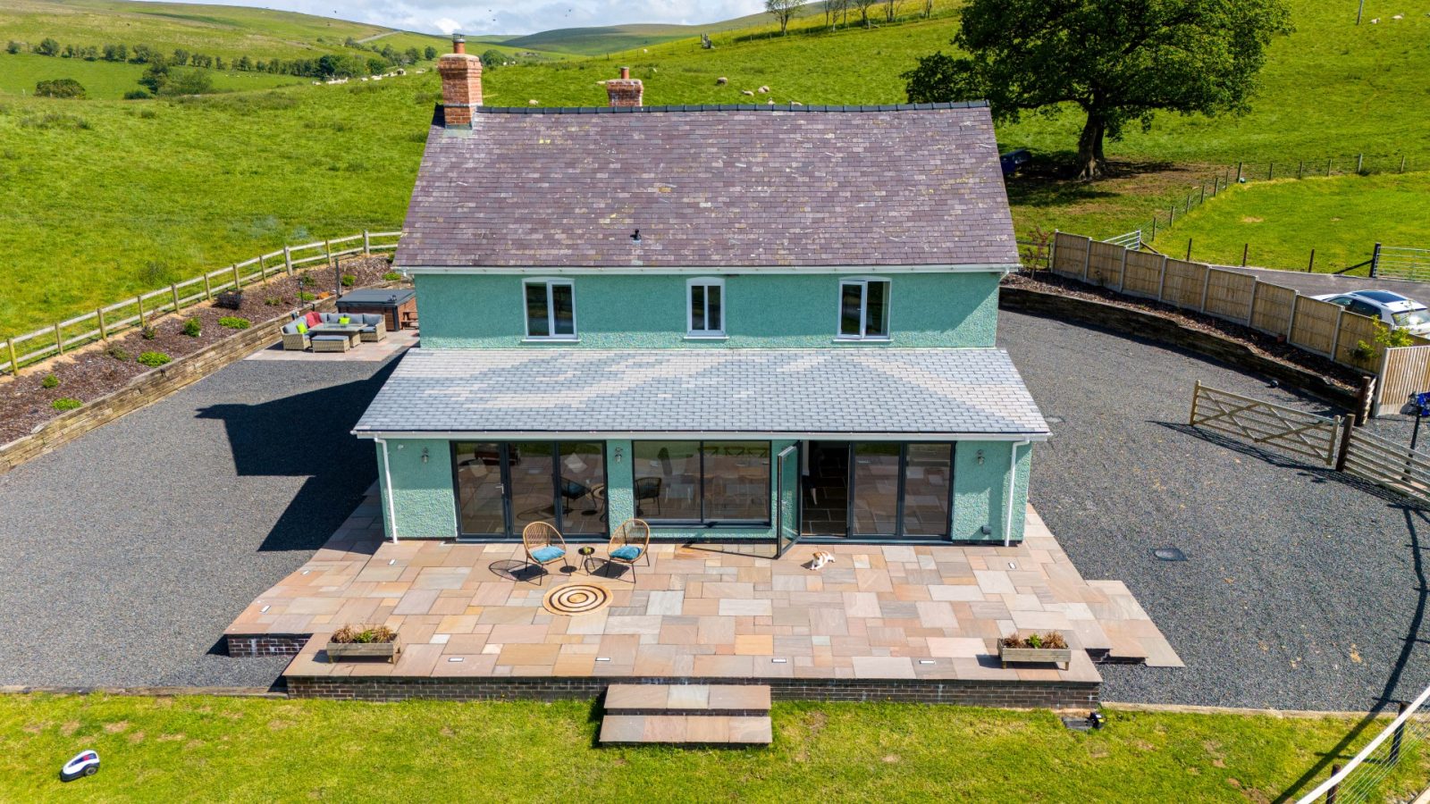 The Waen Farm House is an undetached two-story home with a green exterior. It features a spacious patio area complete with outdoor seating and a table. Nestled in a rural, hilly area surrounded by lush green fields, it boasts stone steps leading to the patio and a picturesque tree in the background.