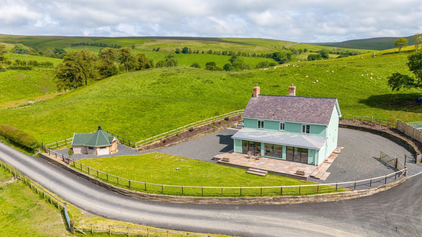 A large, light green house with a paved driveway and surrounding grassy hills. A small, round outbuilding is nearby. A winding road runs in front of the property, and sheep graze on the distant hills under a partly cloudy sky at Waen Farm House.