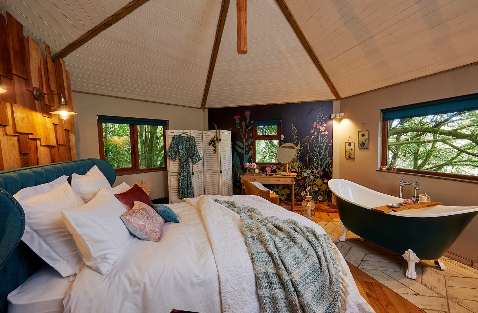 A cosy bedroom with wooden ceiling and walls features a large bed with white linens and green accents. A freestanding bathtub with a wooden tray stands nearby. A wardrobe screen and dressing table complement the serene space, with windows showcasing lush greenery outside—truly a dreamy Devon stay.