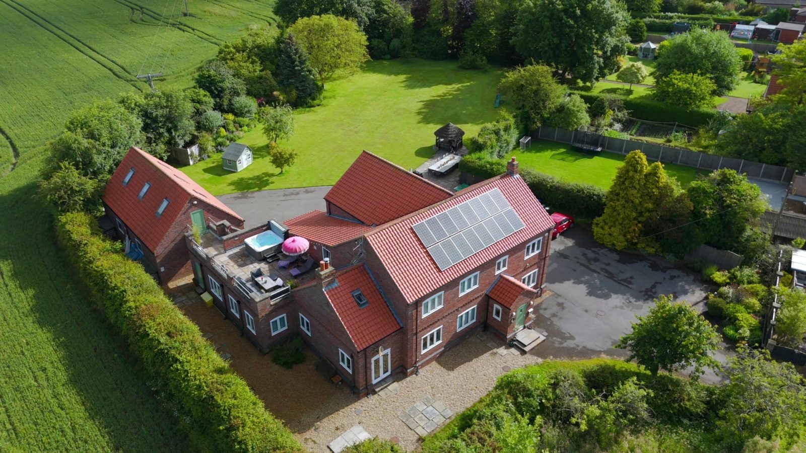 Aerial view of a large brick house with red-tiled roofs and solar panels in West Acre. The property boasts a spacious garden encircled by lush greenery and a winding driveway. Adjacent structures include a garage or outbuilding.