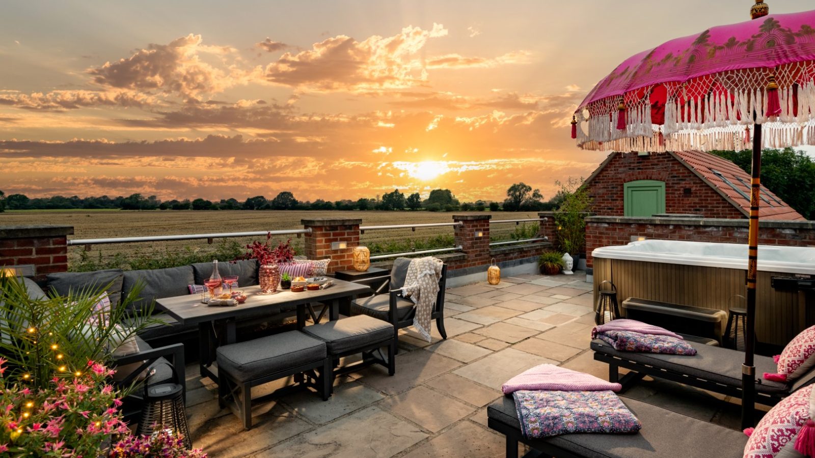 A spacious rooftop terrace at sunset in West Acre features a dining table with dark chairs, colorful cushions, flowers, and a pink parasol with tassels beside the inviting hot tub. The sky is filled with dramatic clouds and sunlight, offering a breathtaking view of the vast field in the distance.