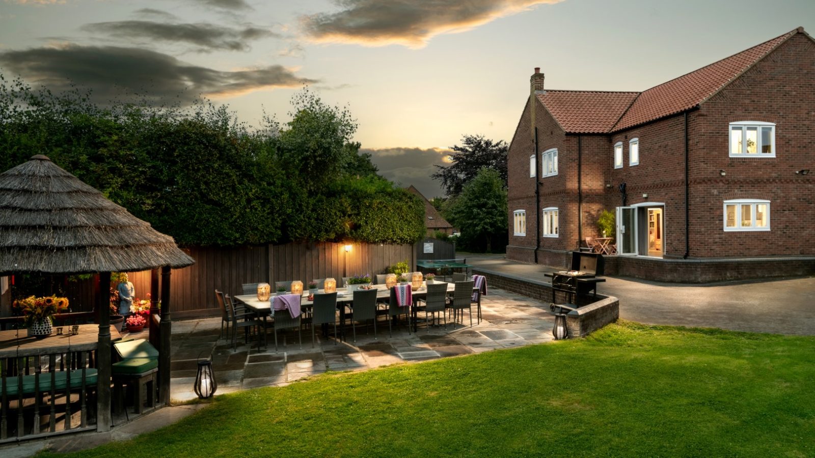 As dusk settles over West Acre, the spacious backyard reveals a long dining table set for a meal on the stone patio. A thatched-roof gazebo sits to the left, while a grand red-brick house stands in the background, surrounded by well-maintained lawns and trees.