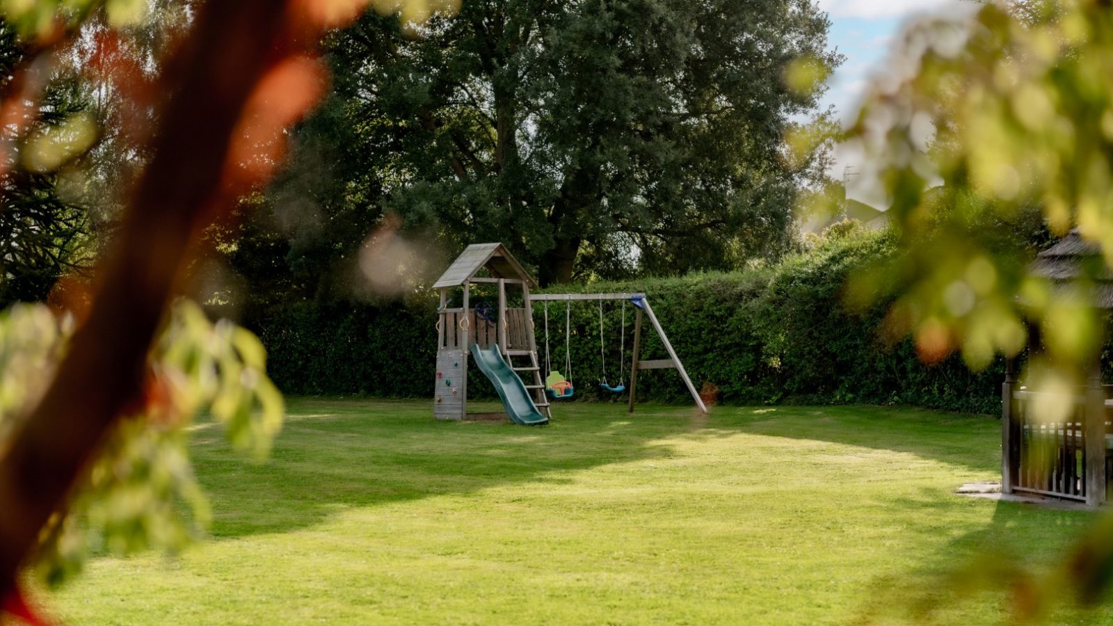 A playground in a grassy backyard in West Acre features a swing set and a slide. Tall green hedges border the area, with trees in the background. Blurred leaves are visible in the foreground, suggesting a sunny day.