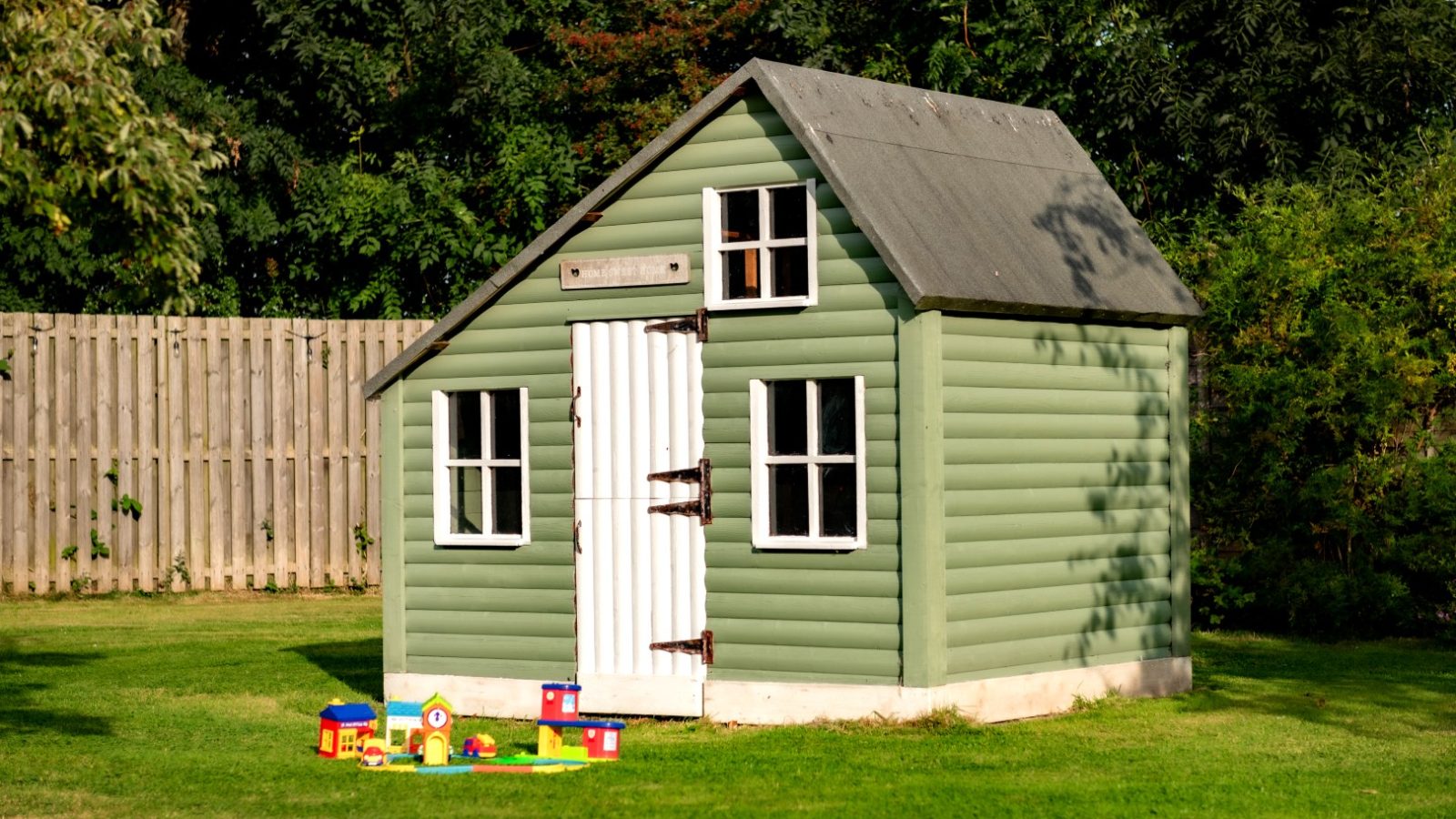 A small, green wooden playhouse with white trim and a gray roof stands on the lush West Acre lawn. In front of the playhouse, colorful toy blocks and a toy house set await eager hands. Trees and a wooden fence create a charming backdrop.