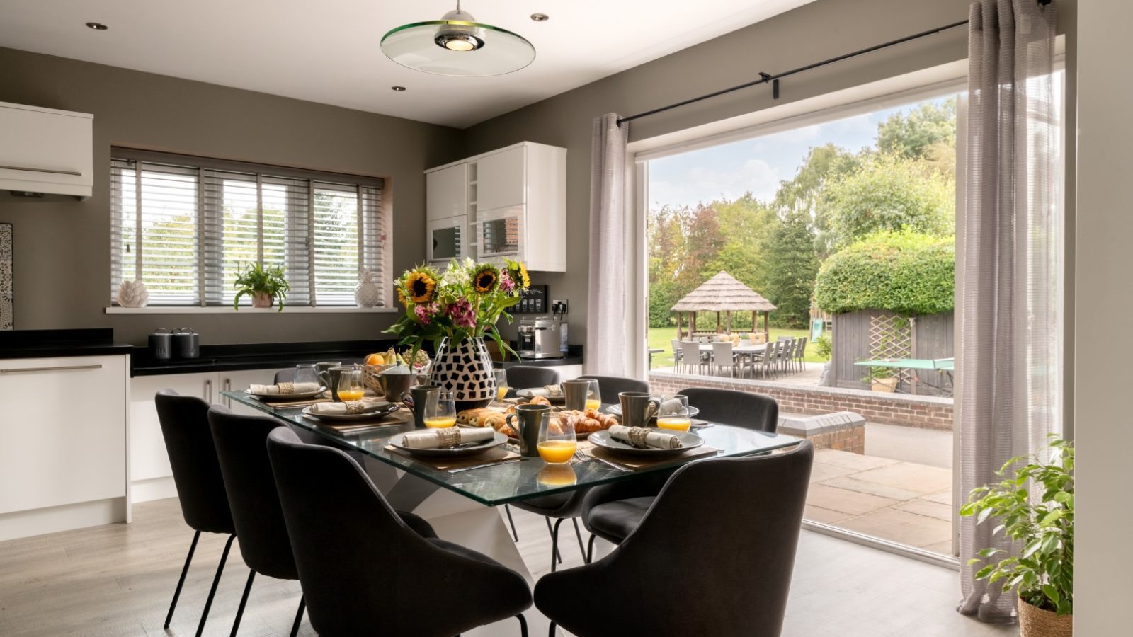 A modern dining room in West Acre showcases a glass table set for a meal, flanked by sleek black chairs and sunflowers in a vase. Through the open patio door, there's a glimpse of the lush garden with outdoor seating surrounded by vibrant greenery.