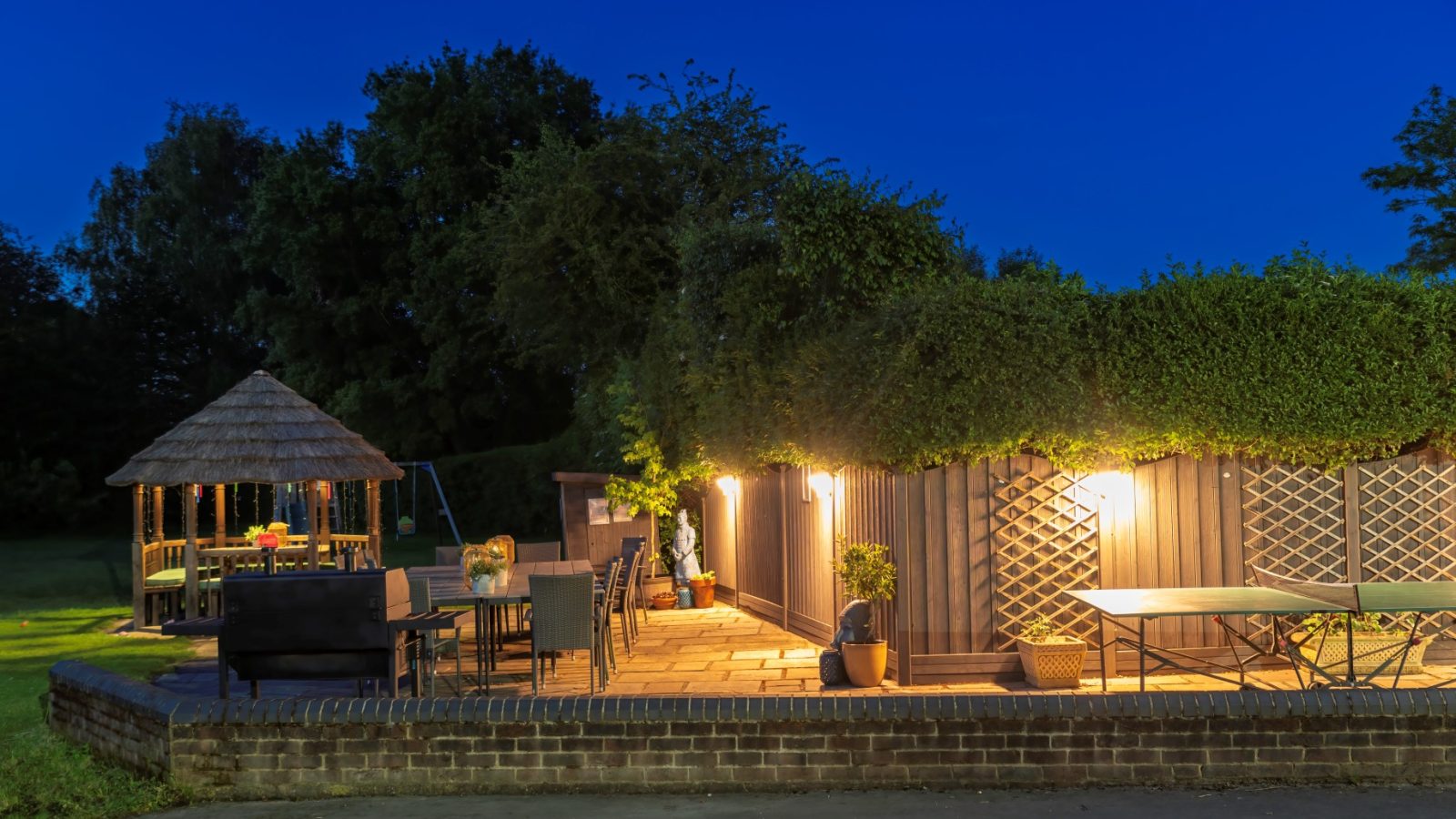 A patio at dusk with a lit outdoor seating area at West Acre, surrounded by lush greenery. A thatched gazebo is visible in the background, and a ping pong table is in the foreground. Warm lights create a cozy, inviting atmosphere.