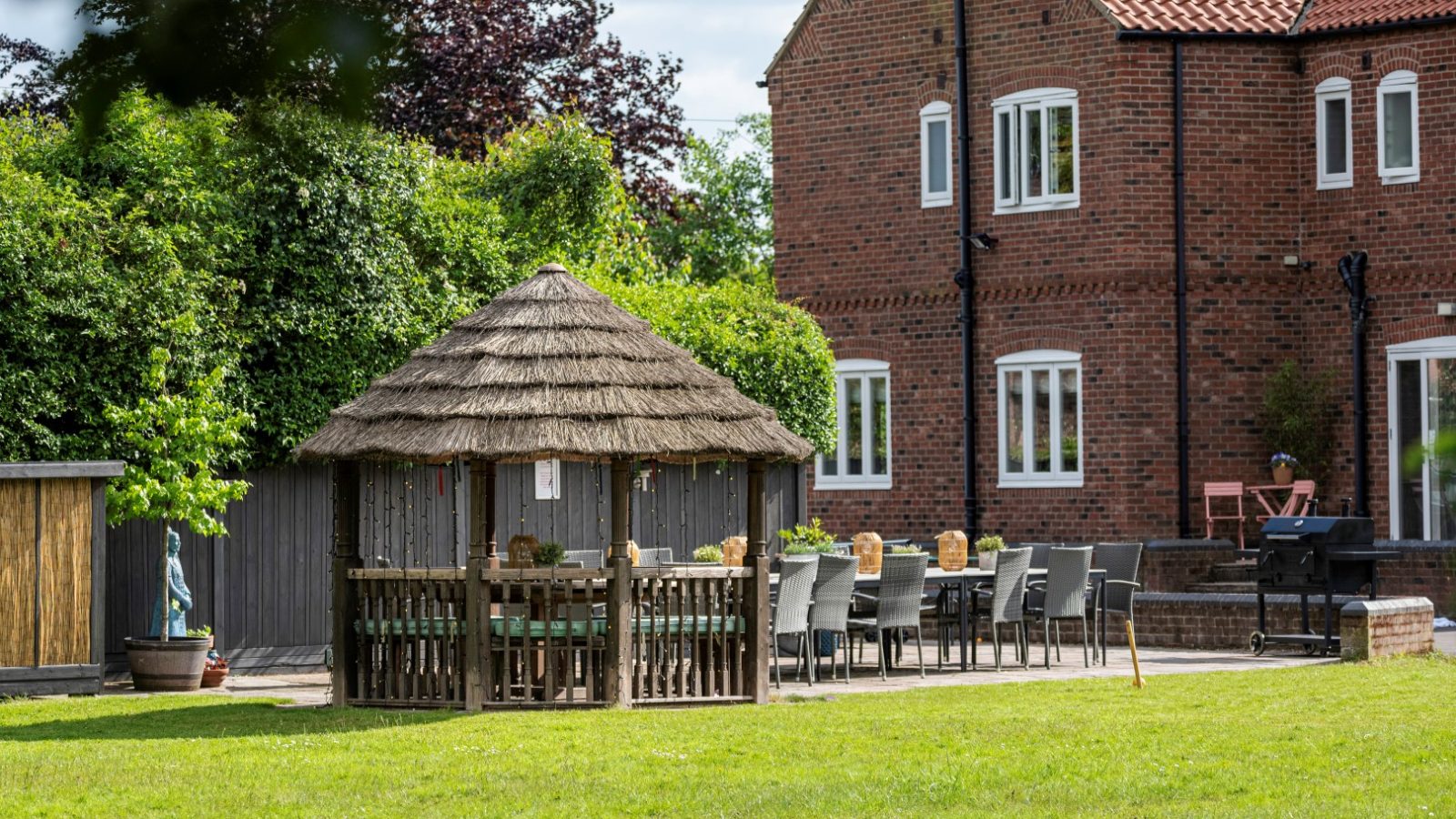 A red-brick house in West Acre stands next to a large yard featuring a thatched-roof gazebo with chairs. Outdoor dining tables are set up nearby. Lush green grass and trees frame the background under a partly cloudy sky.