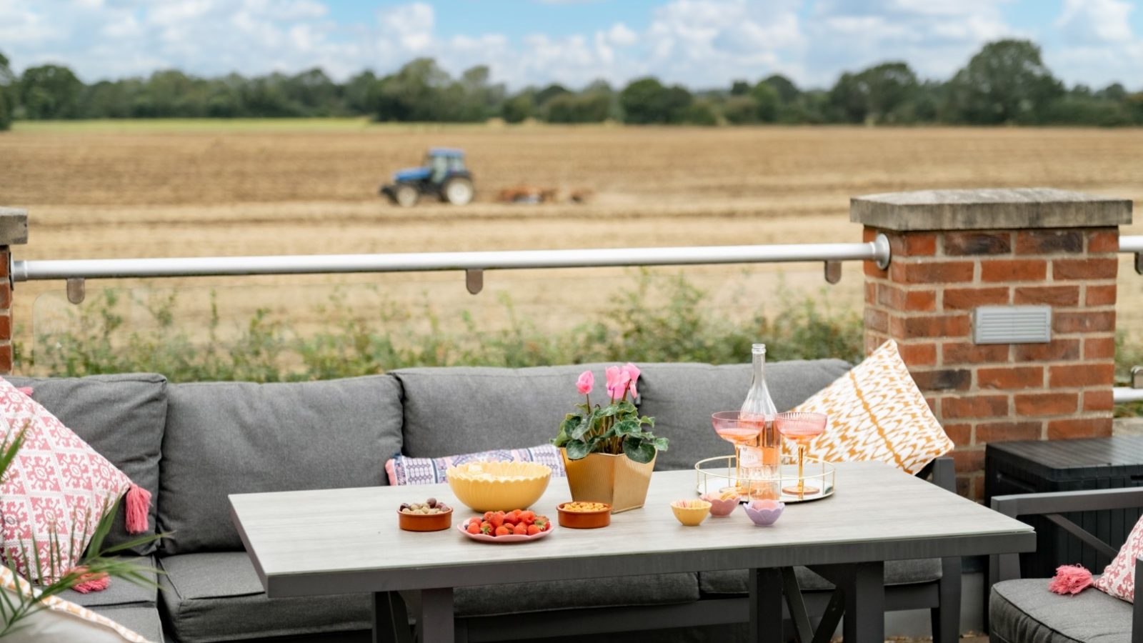 The outdoor patio at West Acre boasts a gray couch and table set, adorned with pink cushions. A delightful array of snacks and drinks awaits while, in the background, a field hosts a tractor under the cloudy sky.