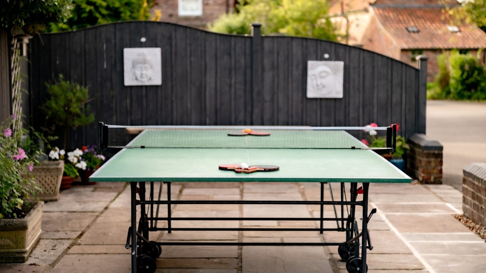 A green outdoor ping pong table is set up on a stone patio with paddles strewn across it. In the background, a black wooden fence adorned with two paper drawings hints at the charming community of West Acre, surrounded by lush greenery and two brick buildings.