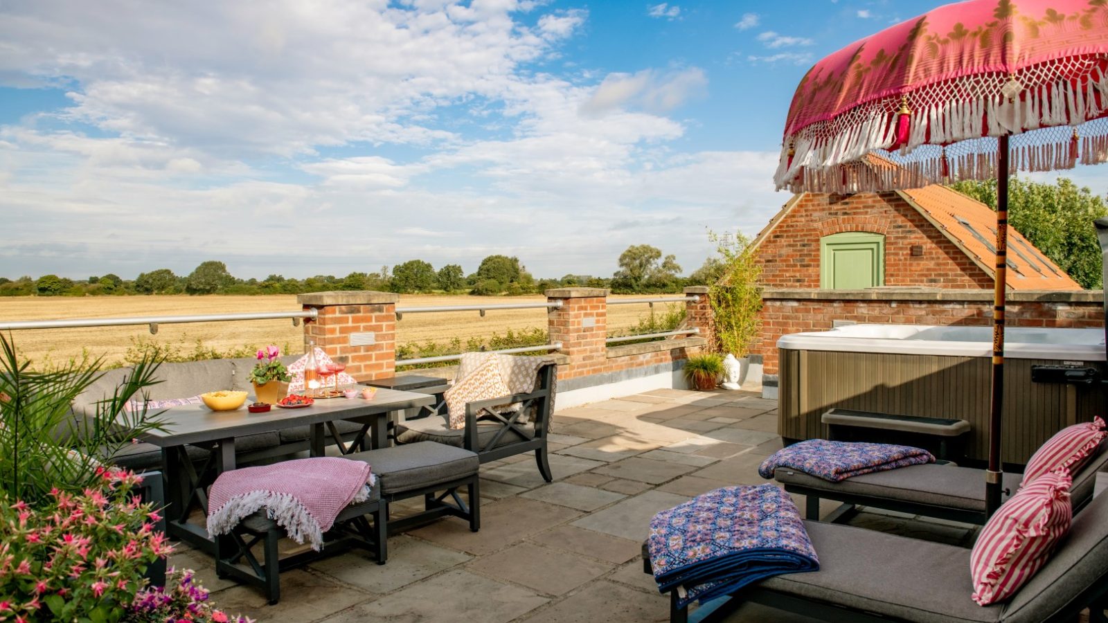 A sunny patio in West Acre features lounge chairs, a dining table with snacks, and a vibrant pink parasol. Brick walls enclose the space with a hot tub nearby. The view offers a green field and scattered trees beneath a blue, partly cloudy sky.