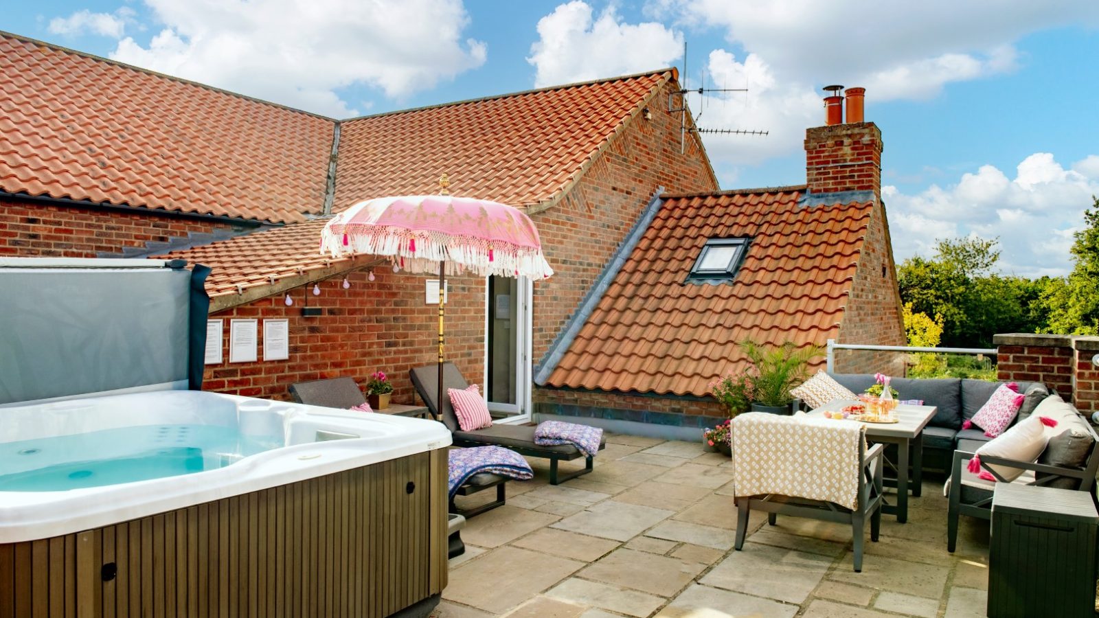 The rooftop patio at West Acre boasts a hot tub, sun loungers with cushions, and a pink fringed umbrella, all set against charming brick walls. A table is elegantly set for dining under partly cloudy skies.