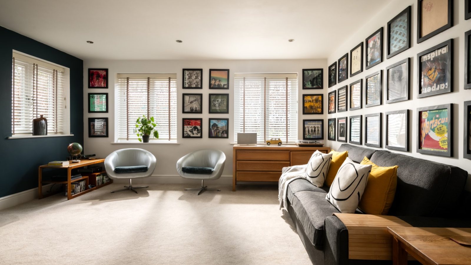 A well-lit West Acre living room with a wall covered in framed art prints. There's a dark sofa with yellow and white pillows, a wooden side table, and two modern gray armchairs by the window. A wooden dresser and bookshelf sit under the window, while a plant adds a touch of greenery.