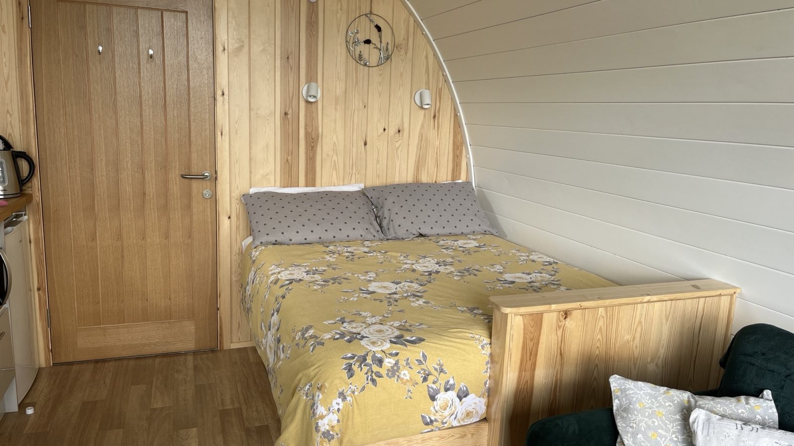 A cozy wooden cabin interior reminiscent of Knighton, featuring a neatly made bed with a yellow floral bedspread and gray pillows. The walls are paneled with light wood and white sections. There's a small kitchenette on the left and a green couch with decorative pillows on the right—classic Wigwam Holidays charm.