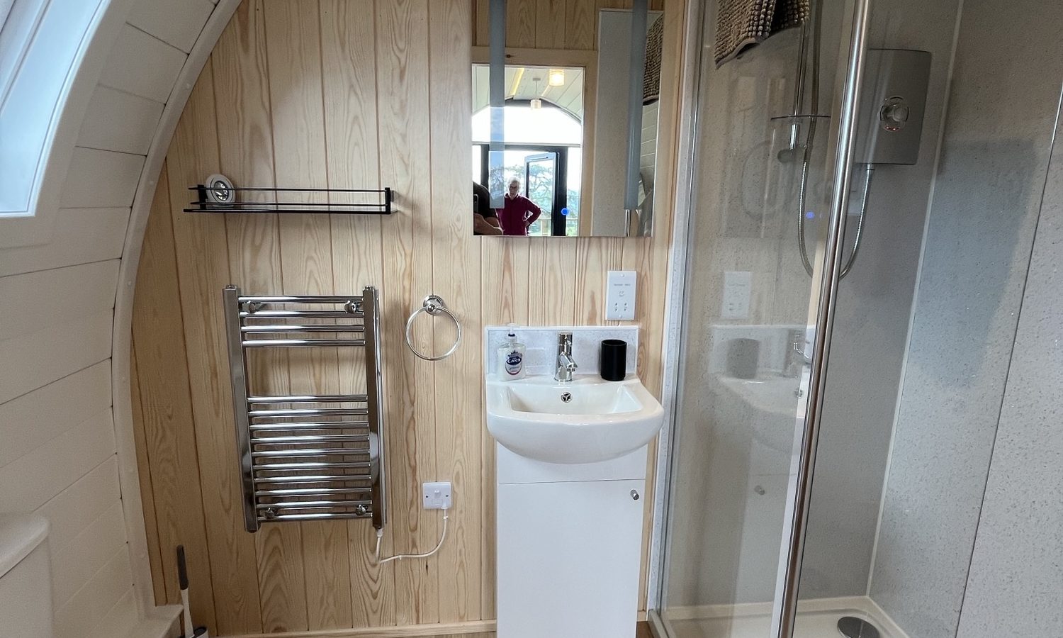 A modern, compact bathroom with light wooden walls and flooring, perfect for those staying at Wigwam Holidays in Knighton. It includes a toilet, a towel rack, a small white sink with a mirror above it, and a glass-enclosed shower. There is a window on the left wall, allowing natural light to enter the space.