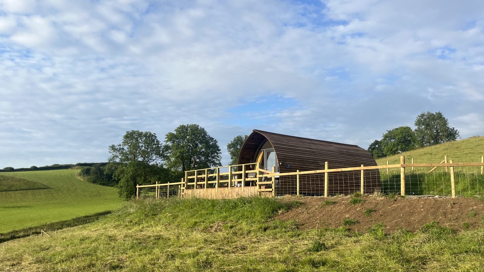 A small, curved wooden cabin, offered by Wigwam Holidays, sits on a grassy hill surrounded by a wooden fence. The sky above Knighton is partly cloudy, and rolling green hills extend into the distance, creating a peaceful countryside scene.