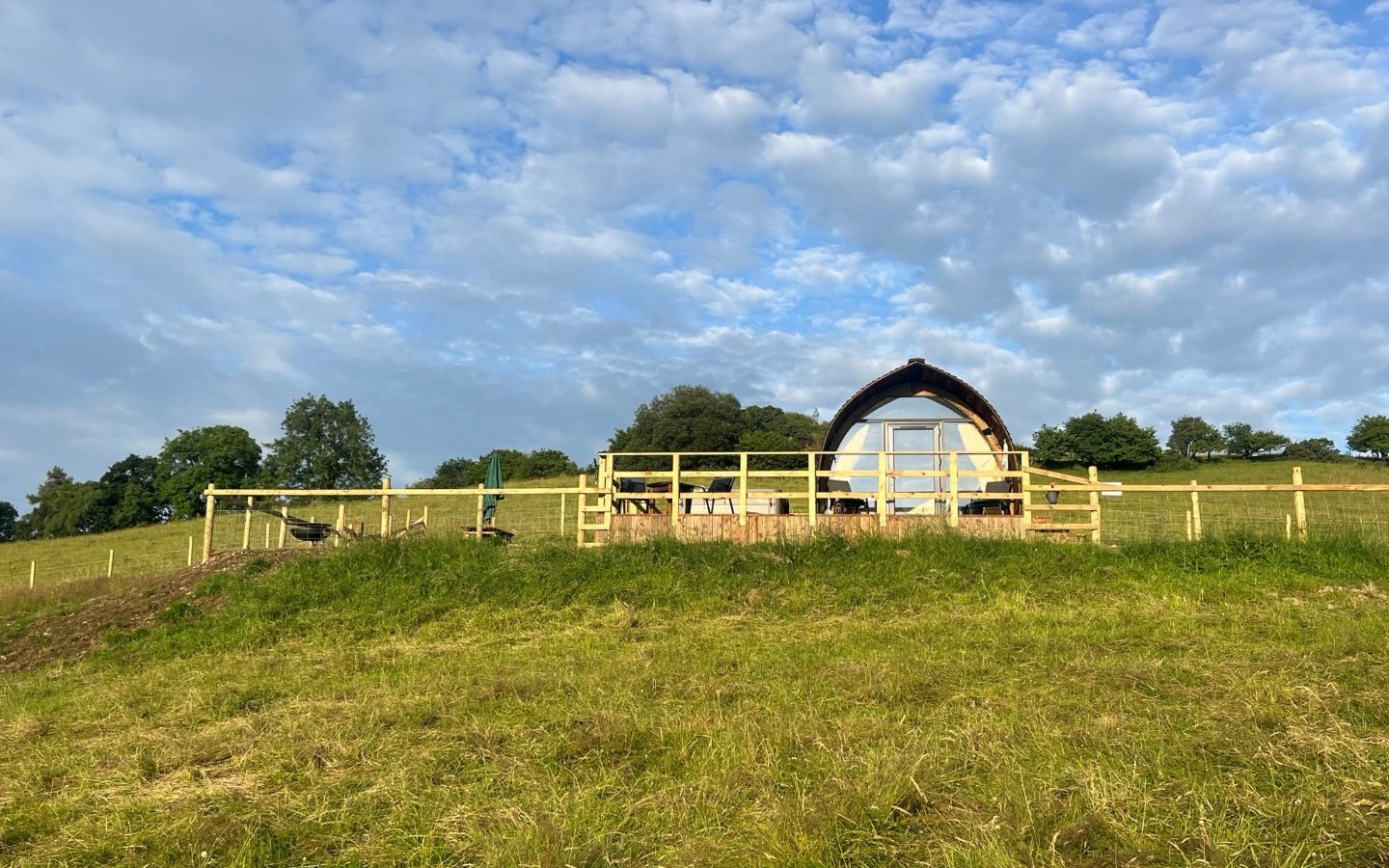 A small, curved-roof cabin with a partially transparent front stands on a grassy hill surrounded by wooden fencing. The sky above is blue with scattered clouds, and the background features rolling green hills and trees, reminiscent of Wigwam Holidays in Knighton.