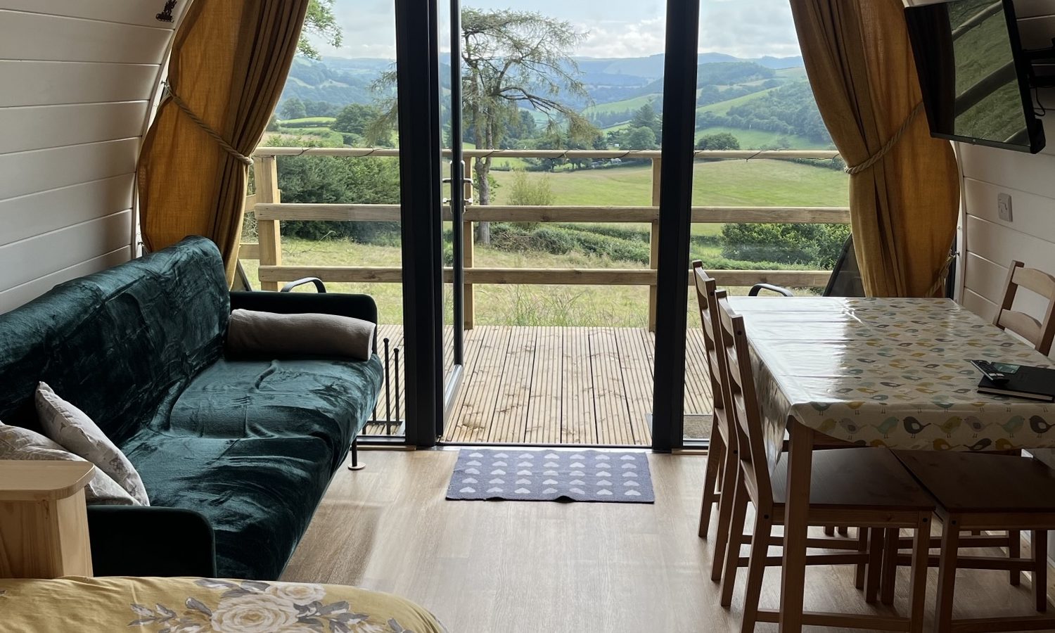         A cozy cabin interior featuring a green sofa, a dining table with chairs, and a bed with a flower-patterned quilt. Large glass doors open to a deck offering scenic views of rolling green hills under a partly cloudy sky in Knighton. Yellow curtains frame the doors, reminiscent of Wigwam Holidays charm.