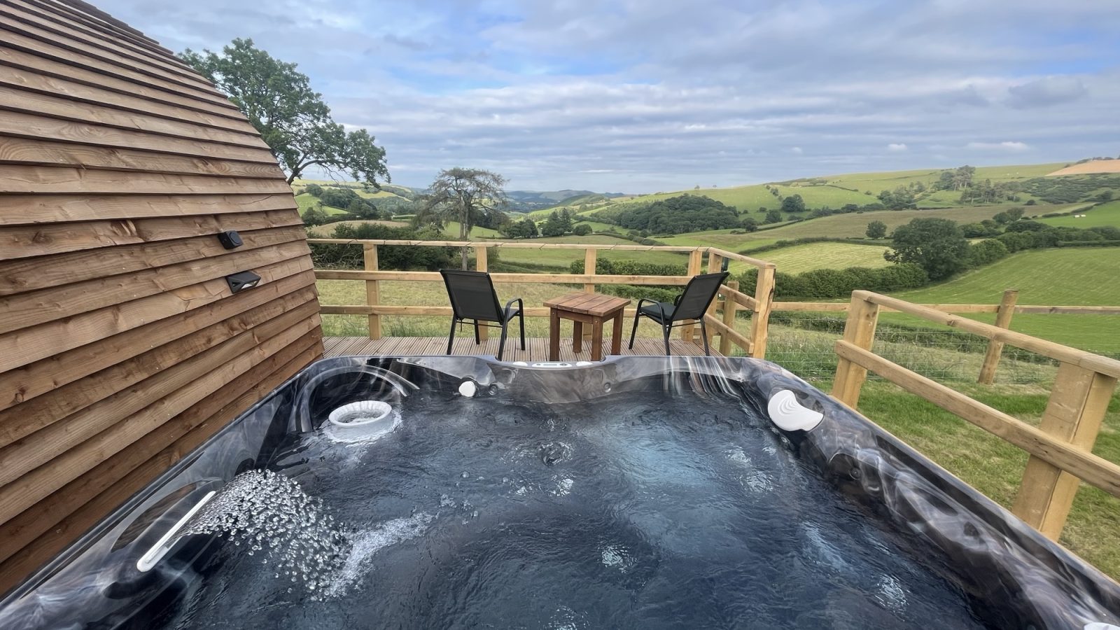 A hot tub sits on a wooden deck with bubbling water, adjacent to a wooden building. Beyond, two black chairs and a small wooden table invite relaxation against the scenic backdrop of rolling green hills under a partly cloudy sky, making this the perfect Wigwam Holidays escape in Knighton.