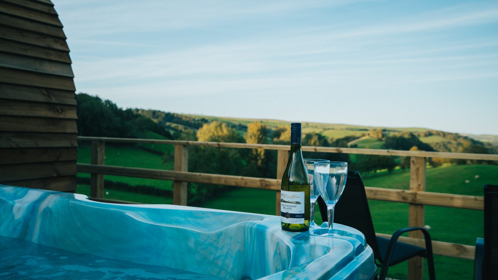 A scenic countryside view with rolling green hills under a blue sky near Knighton. In the foreground, there's a hot tub with a bottle of white wine and two wine glasses placed on its edge. A wooden fence and outdoor seating from Wigwam Holidays are visible in the background.