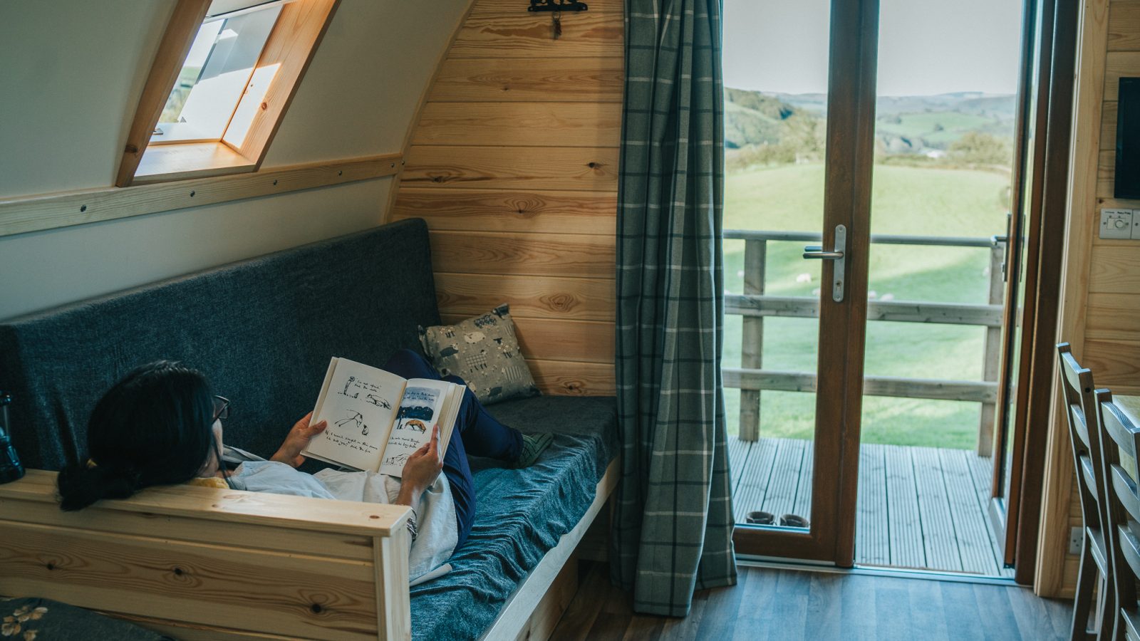 A person reclines on a couch inside a cozy cabin with wooden walls, reading a book. Sunlight streams in through a skylight and a glass door, which opens to a scenic view of grassy hills and a wooden deck outside. A curtain is partially drawn beside the door, providing the perfect retreat for Wigwam Holidays near Knighton.