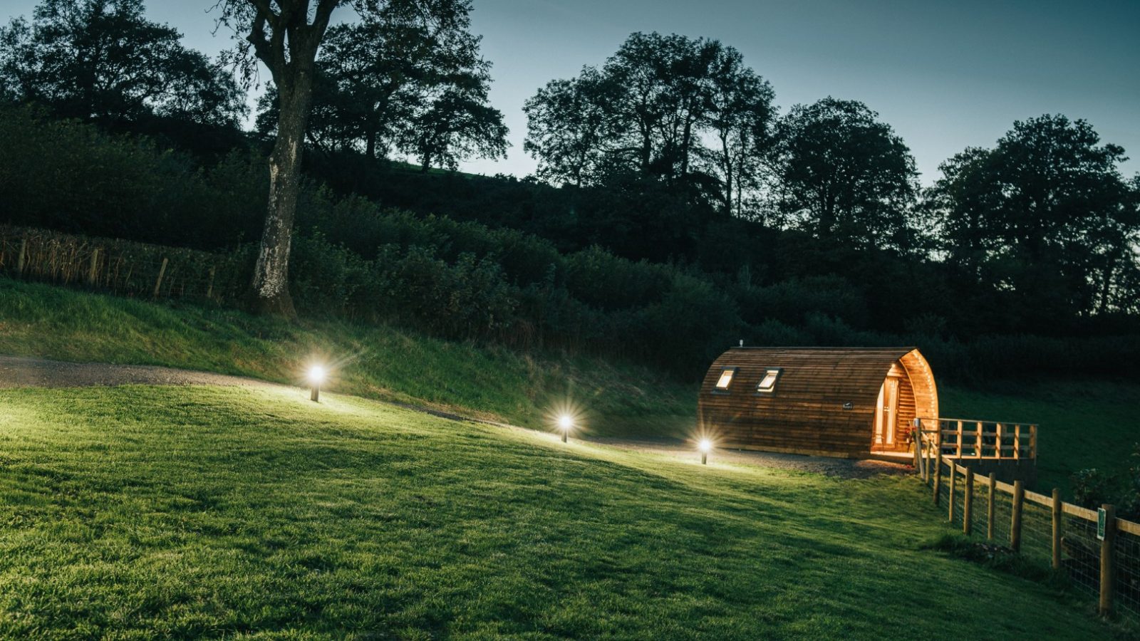 A small, illuminated wooden cabin with an arched roof sits in a grassy field near Knighton at dusk. Trees line the background, and pathway lights lead to the entrance, creating a warm and inviting atmosphere reminiscent of Wigwam Holidays.