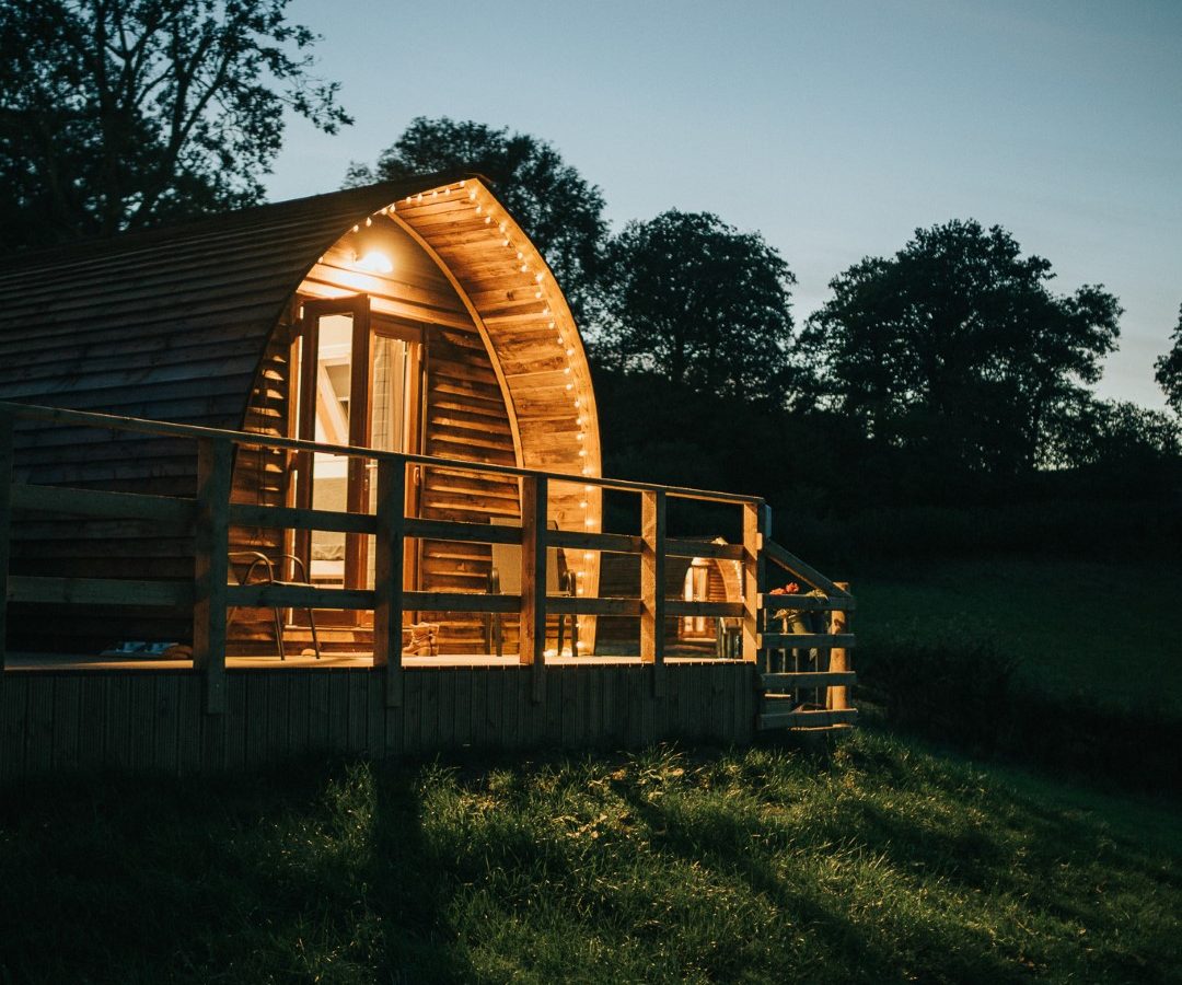 A small, arched wooden cabin with a front porch is illuminated by warm, inviting lights against the dusk sky. Nestled amidst tall trees and a grassy landscape, this tranquil and cozy retreat offers the perfect Wigwam Holidays experience near Knighton.