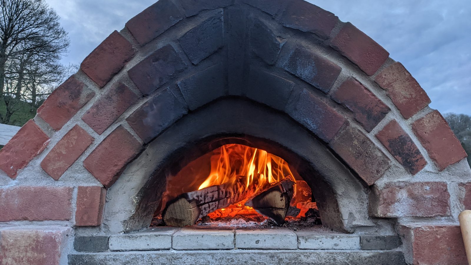 Against a backdrop of a cloudy sky and trees, the outdoor brick pizza oven at Knighton is alive with a wood fire, reminiscent of the warm experiences found on Wigwam Holidays.