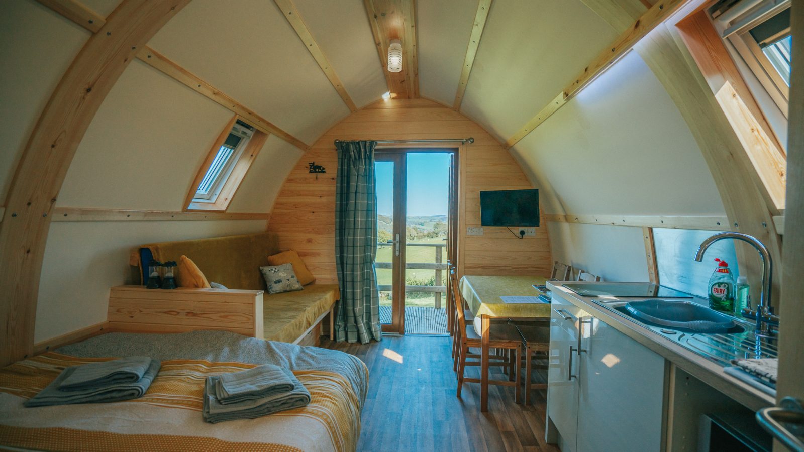 A cozy, wooden interior of a small cabin with a curved ceiling offers all the charm of a Knighton retreat. It features a bed with striped linens, a daybed with cushions, a small kitchen area with a sink, a TV mounted on the wall, and a dining table with chairs. A glass door opens to the scenic beauty typical of Wigwam Holidays.