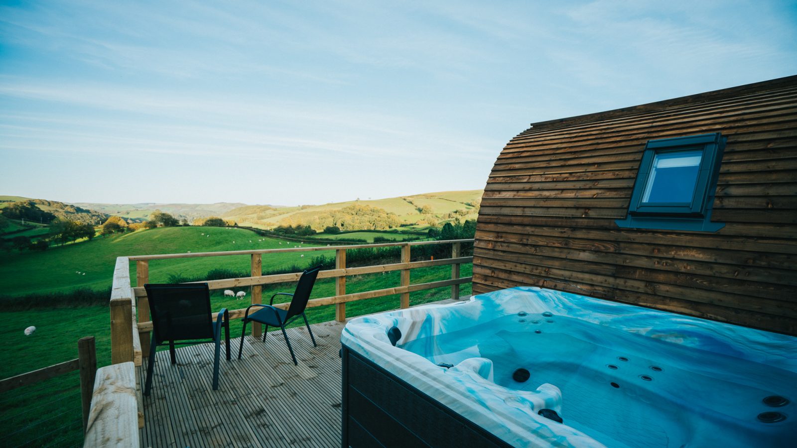 A wooden deck overlooking a scenic, green landscape with hills. On the deck is a hot tub and a small table with two chairs. A wooden cabin wall with a window is to the right. The clear sky and peaceful, remote setting make it an ideal spot for Wigwam Holidays in Knighton.