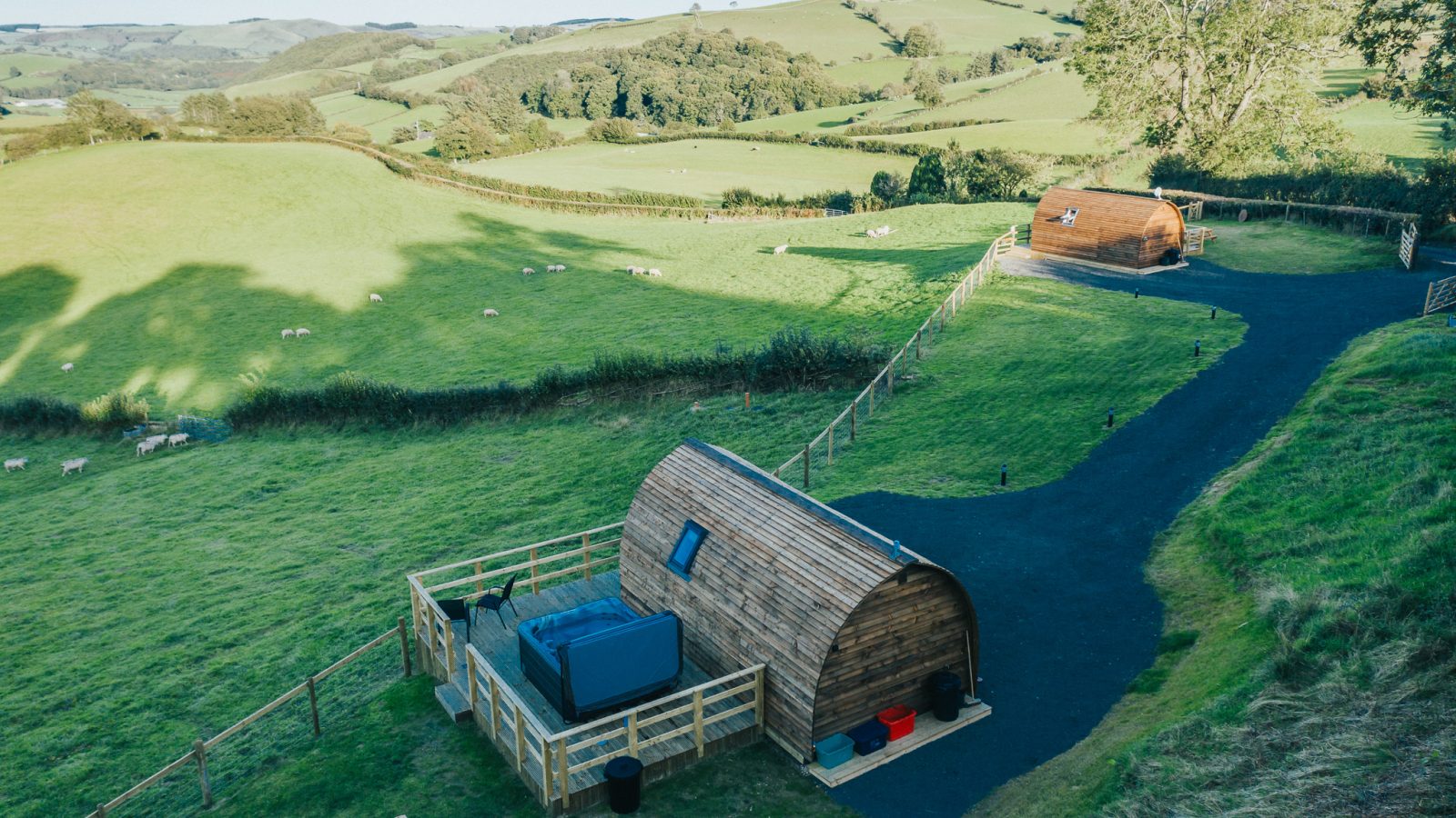 An elevated view of two wooden glamping pods by Wigwam Holidays, set on lush green fields near Knighton. Each pod has its own fenced outdoor area with a picnic table and hot tub. Rolling hills and scattered trees are visible in the background under a clear sky.